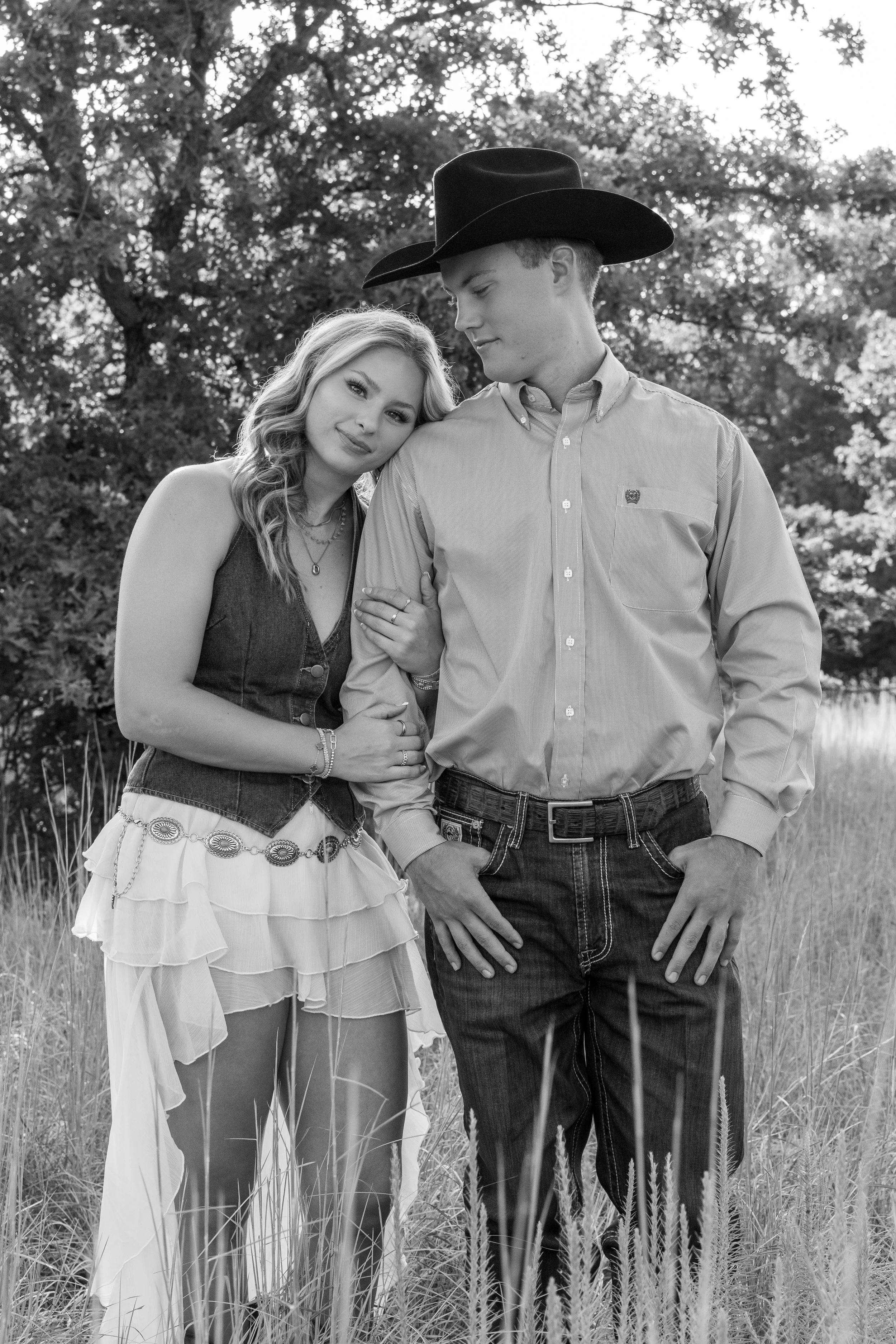 A black and white photo of a young couple standing close together in a grassy field with trees in the background. The woman has wavy hair and wears a sleeveless top and layered skirt, while the man wears a wide-brimmed hat and a striped button-down s