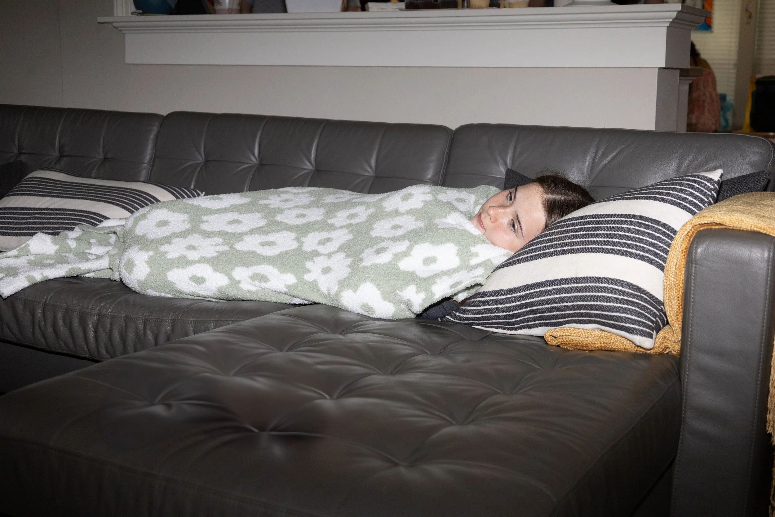 A woman with brown hair lying on a black leather sofa, covered with a green towel with white flower patterns, resting her head on a striped pillow, in a room with a white wall and a yellow towel on the armrest.
