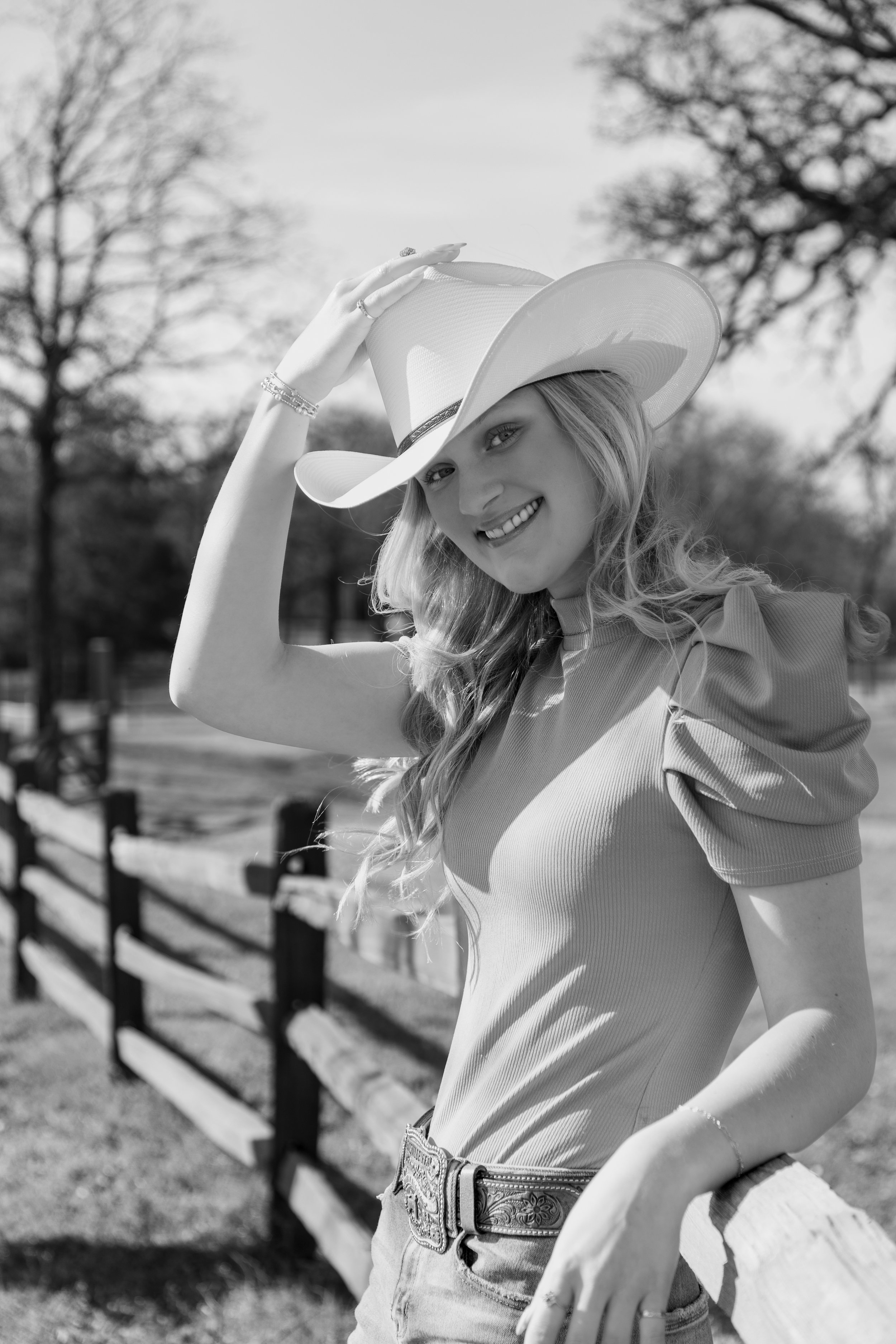 A smiling woman with long hair wearing a wide-brimmed hat, a fitted top with puffy sleeves, jeans, and a decorative belt, standing outdoors next to a wooden fence with trees in the background, in black and white.