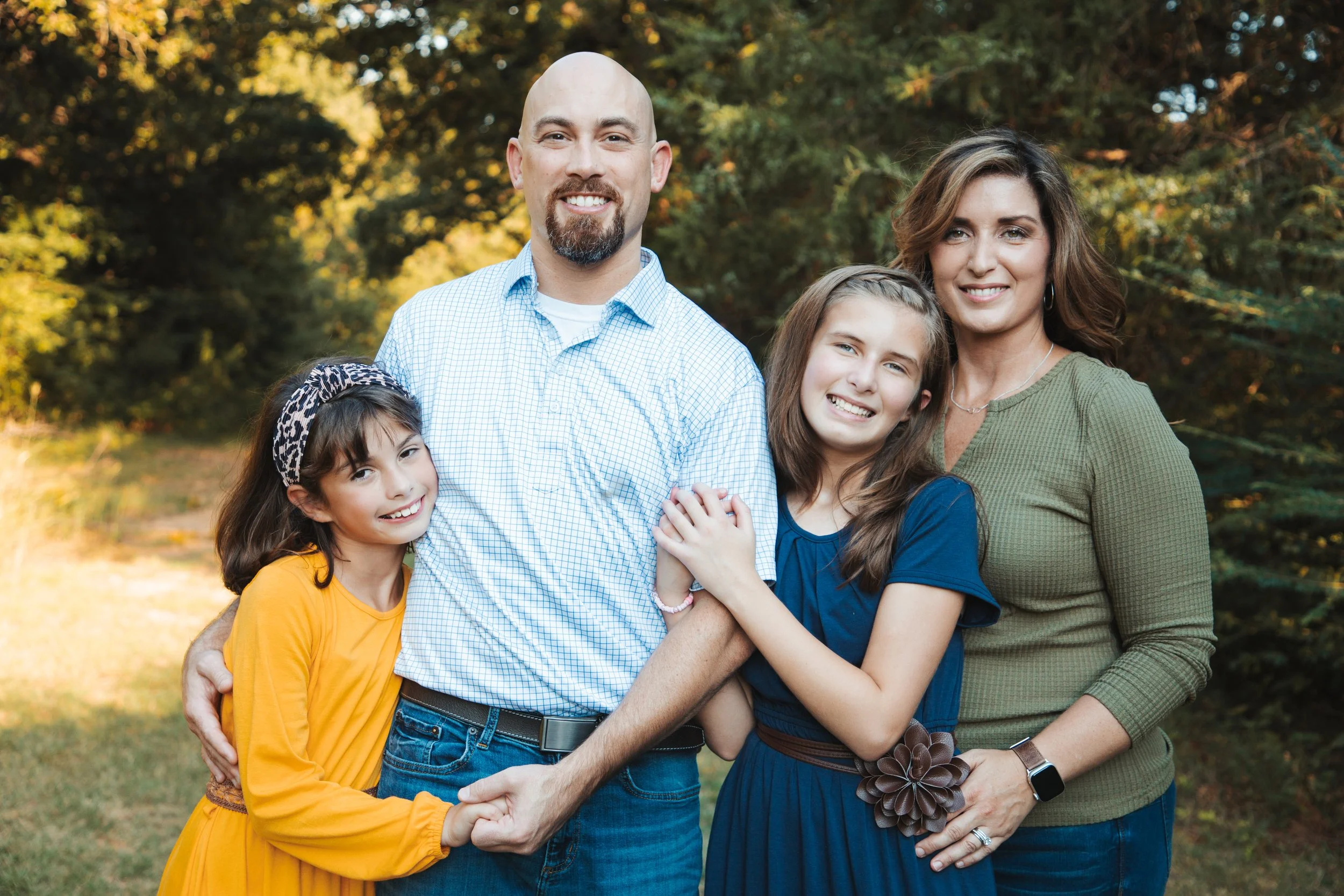 A family of five smiling outdoors in a wooded area during daytime. The father with a beard and bald head, wearing a light blue checkered shirt, is in the center. The mother with wavy brown hair, wearing a green sweater, stands to the right. Two young