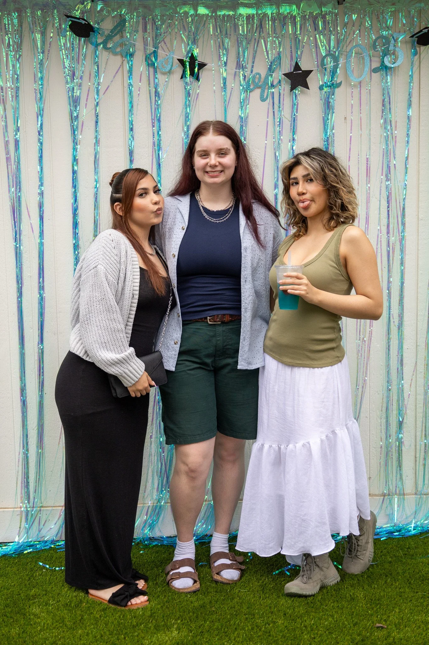 Three young women posing at a party with a colorful, shiny backdrop and a banner that reads 'Graduation 2023' in the background. One is holding a drink, and they are smiling and playful.