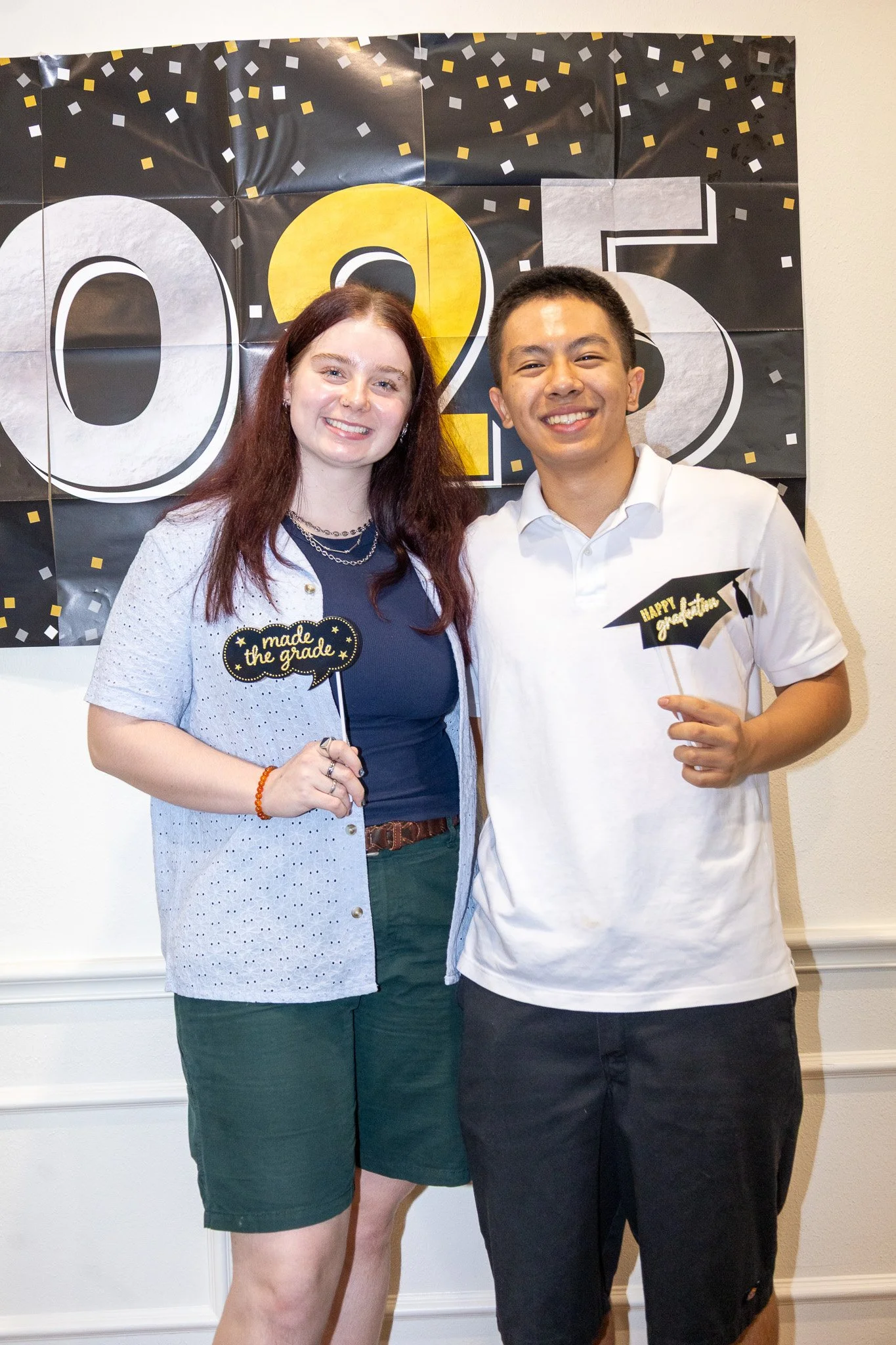 Two young adults, a woman and a man, smiling and posing at a graduation celebration. The woman holds a sign that says 'made the grade,' and the man holds a sign that says 'happy graduation.' A black and yellow banner with '2025' is in the background.