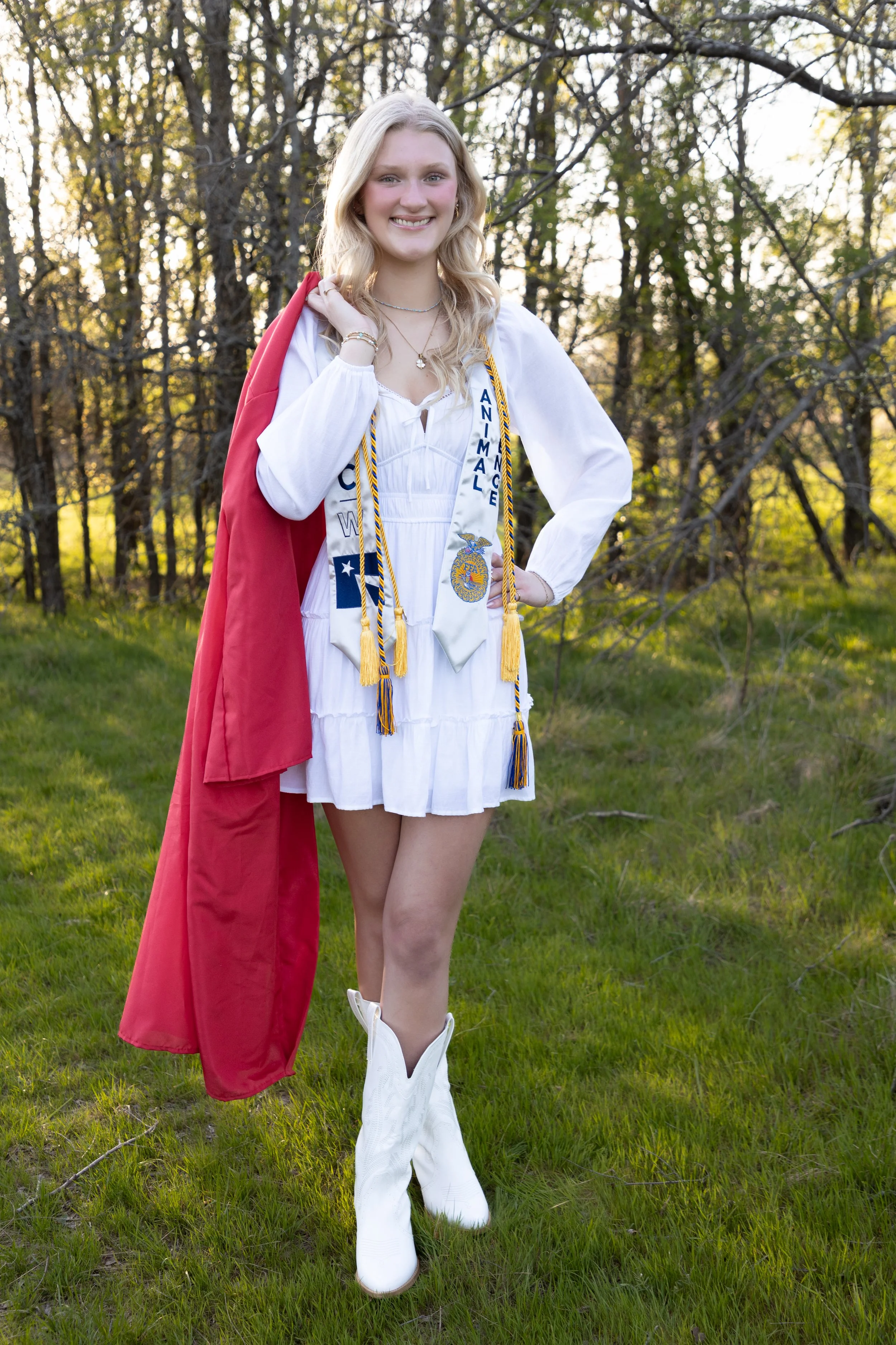 Young woman in a white dress and white cowboy boots, wearing a red graduation gown over her shoulder, standing outdoors on grass with trees in the background during daytime.