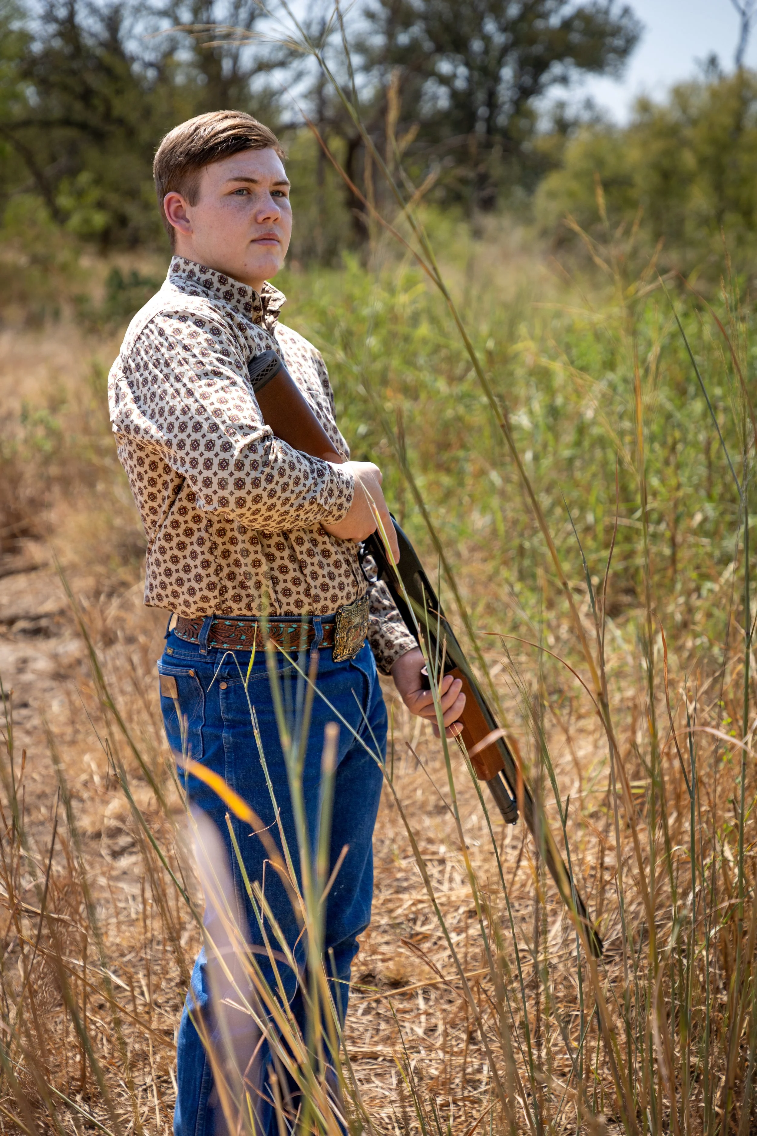 A young man with short brown hair, wearing a patterned button-up shirt and blue jeans, stands in a dry grassy field holding a shotgun. He looks serious and alert, with trees visible in the background.