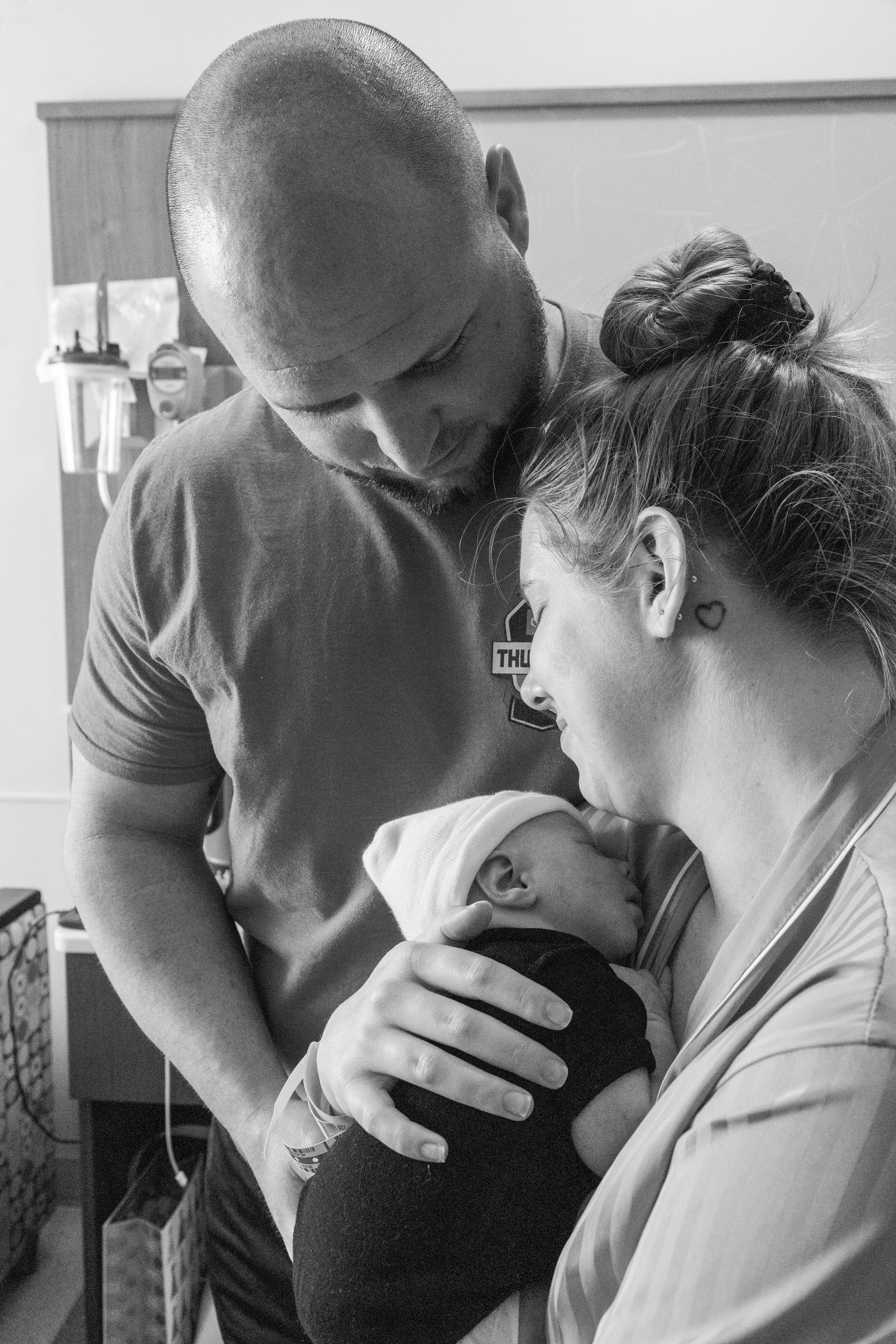 A nurse holding a newborn baby, with a man and woman looking at the baby lovingly in a hospital room.