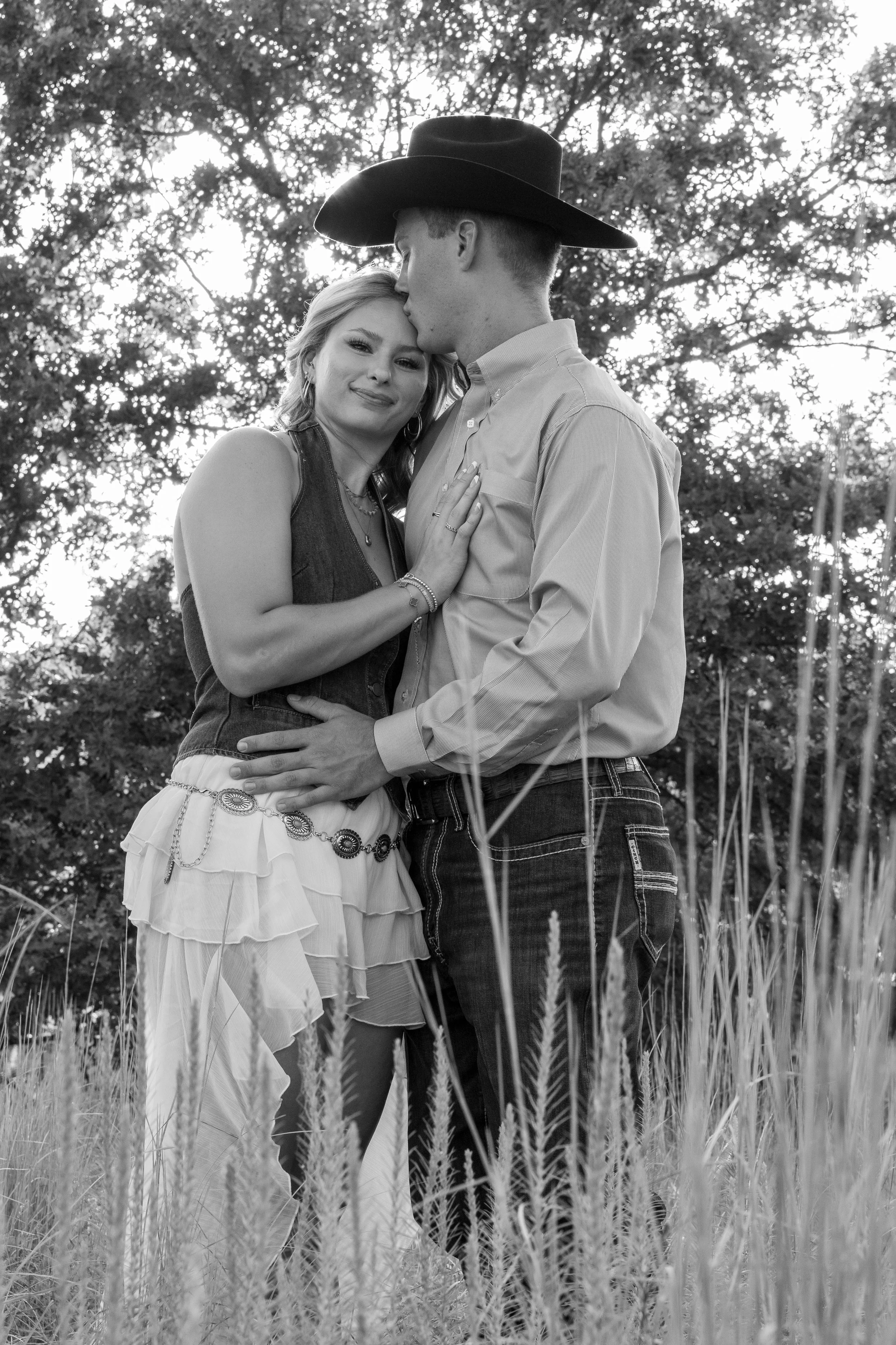 A black and white photo of a couple standing in a field of tall grass, with trees in the background. The woman is wearing a layered skirt and top, and the man is wearing a cowboy hat, button-down shirt, and jeans. The man is kissing the woman's foreh