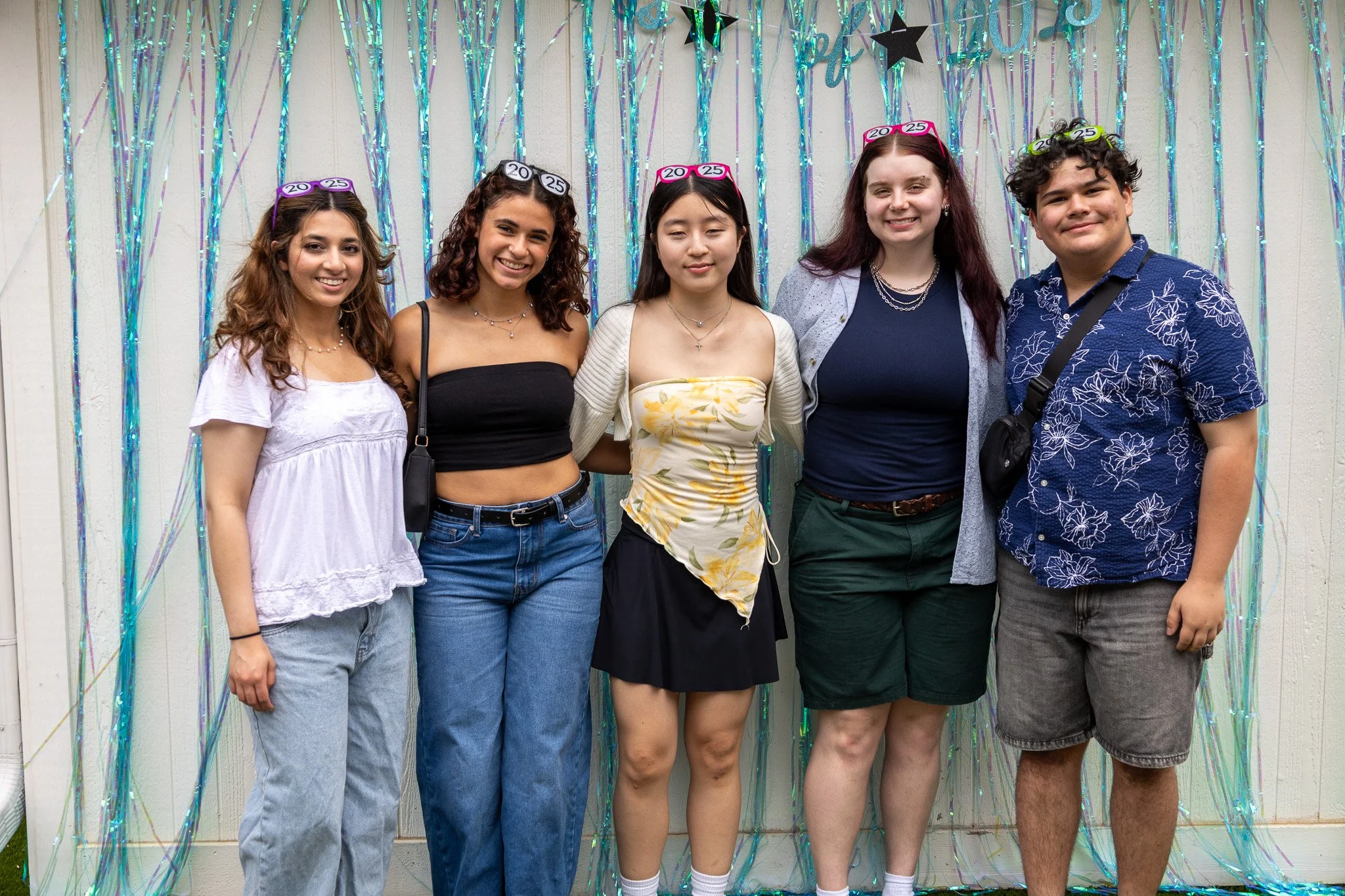 Group of five young people standing together in front of silver and blue streamers, wearing graduation caps celebrating graduation.