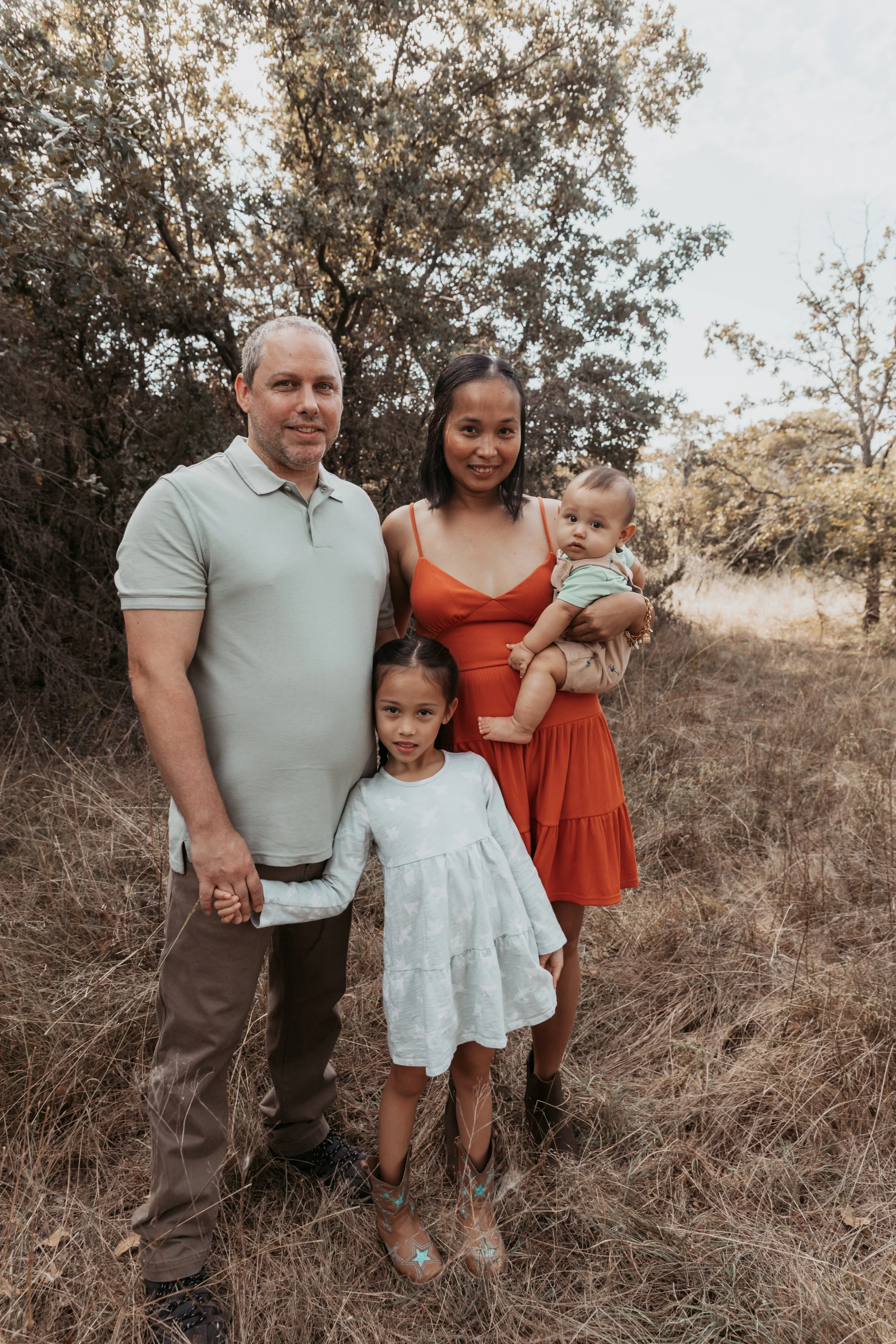 A family of four stands outdoors in a grassy area with trees in the background, smiling at the camera.