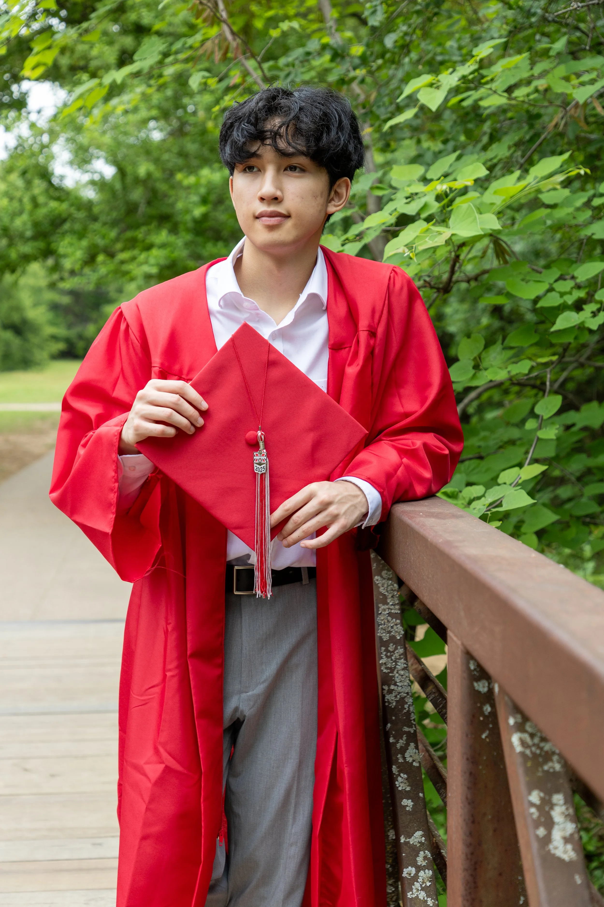 Young man in a white shirt and gray pants wearing a red graduation gown and cap holding his cap with a tassel, standing on a wooden bridge with green trees in the background.