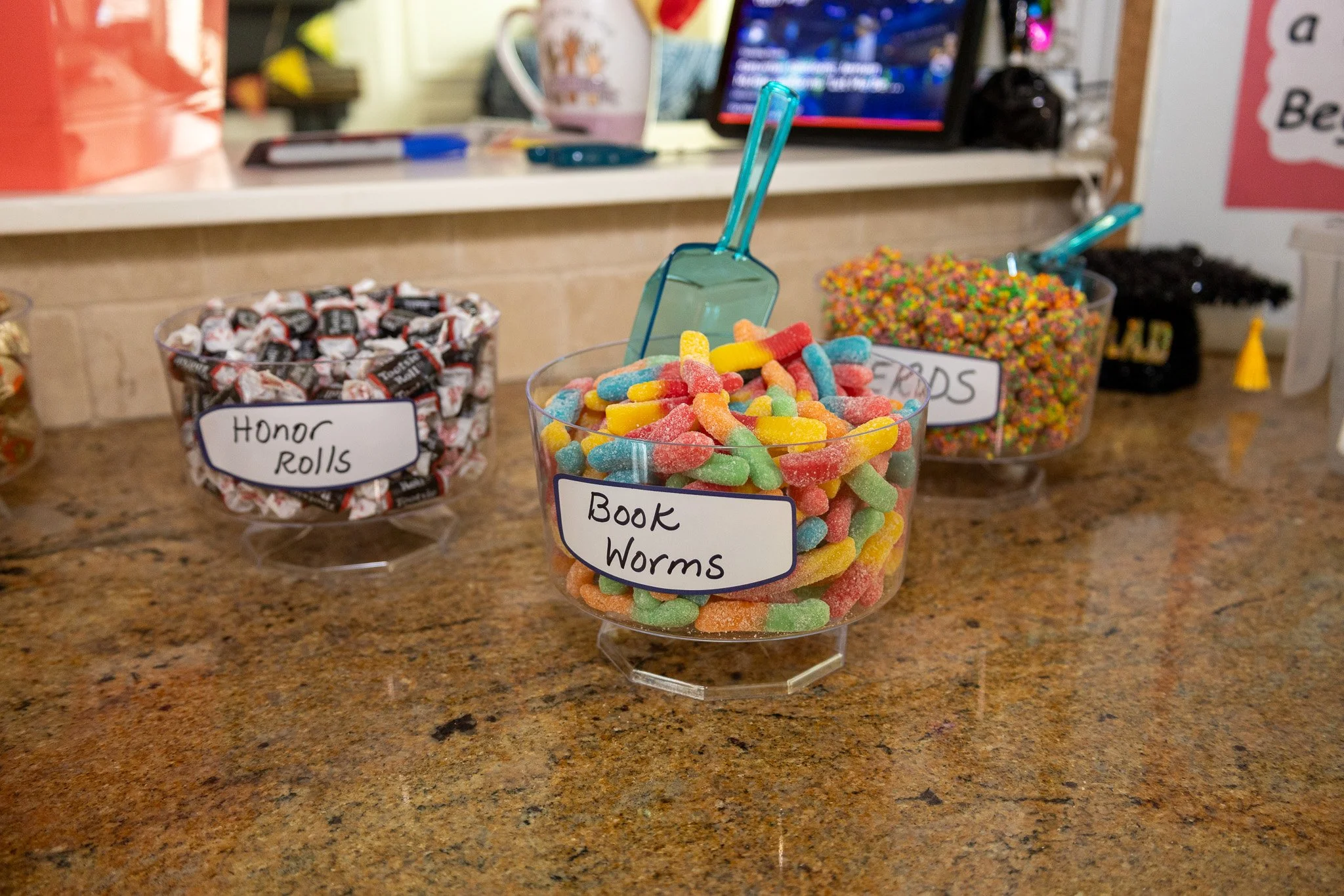 Three containers of assorted candies on a countertop with labels: 'Honor Rolls', 'Book Worms', and 'Friends'. A laptop and other objects are in the background.