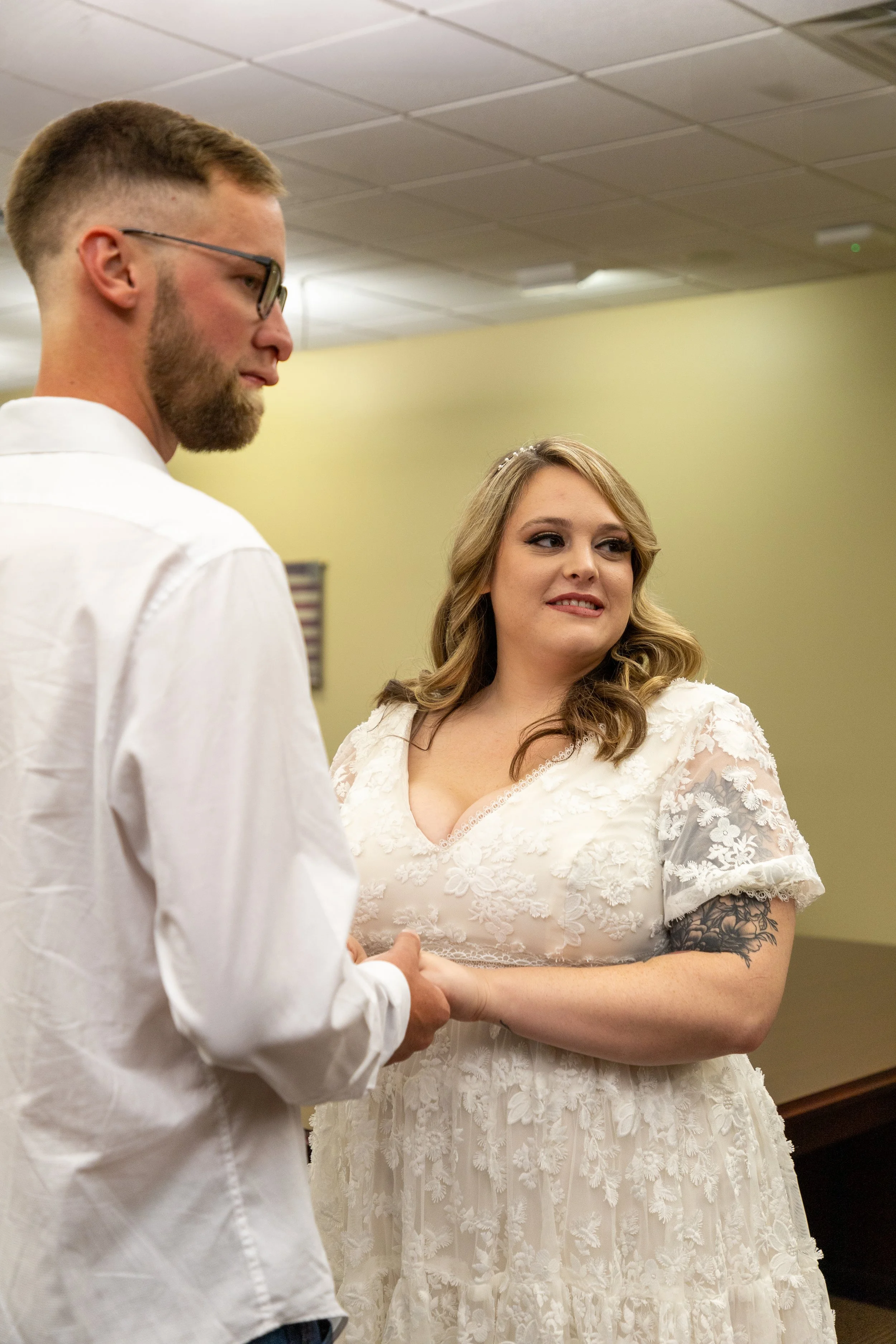 A couple holding hands during a wedding ceremony, with the bride wearing a white lace dress and the groom in a white shirt.