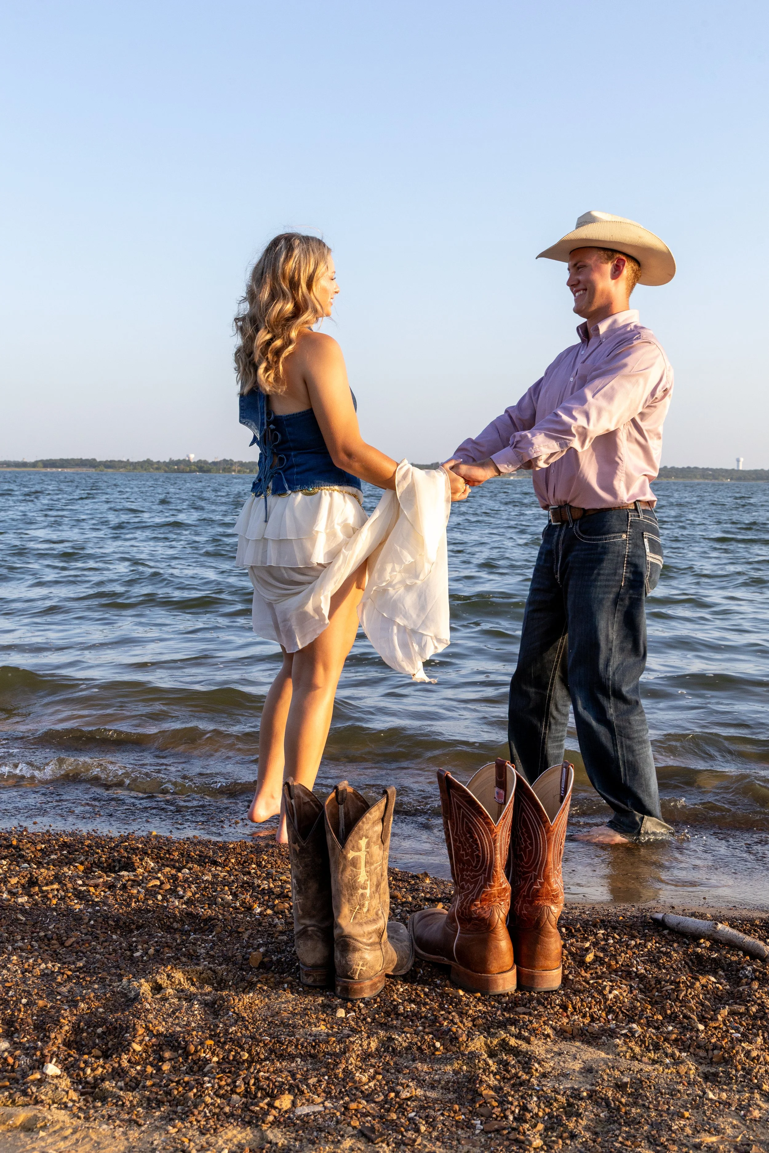 A man and woman holding hands by the water at sunset, with cowboy boots placed on the ground in front of them.