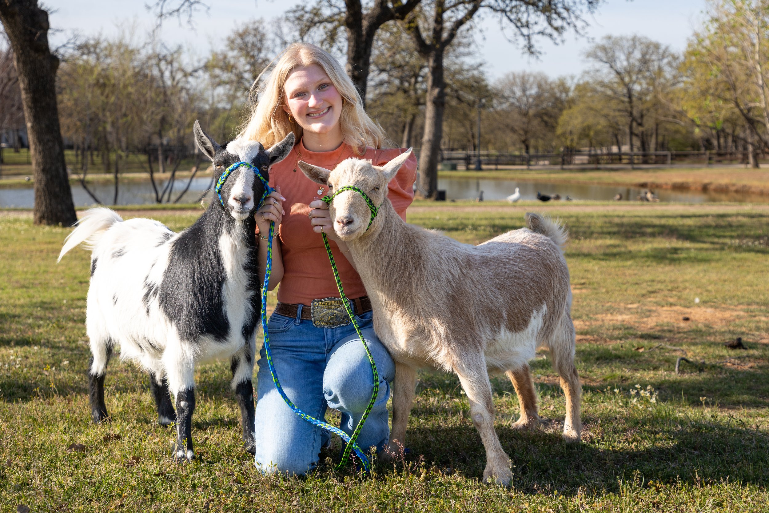 A smiling woman kneeling on grass in a park with two miniature goats, one black and white and the other tan and white, both wearing colorful halters, with a pond and trees in the background on a sunny day.