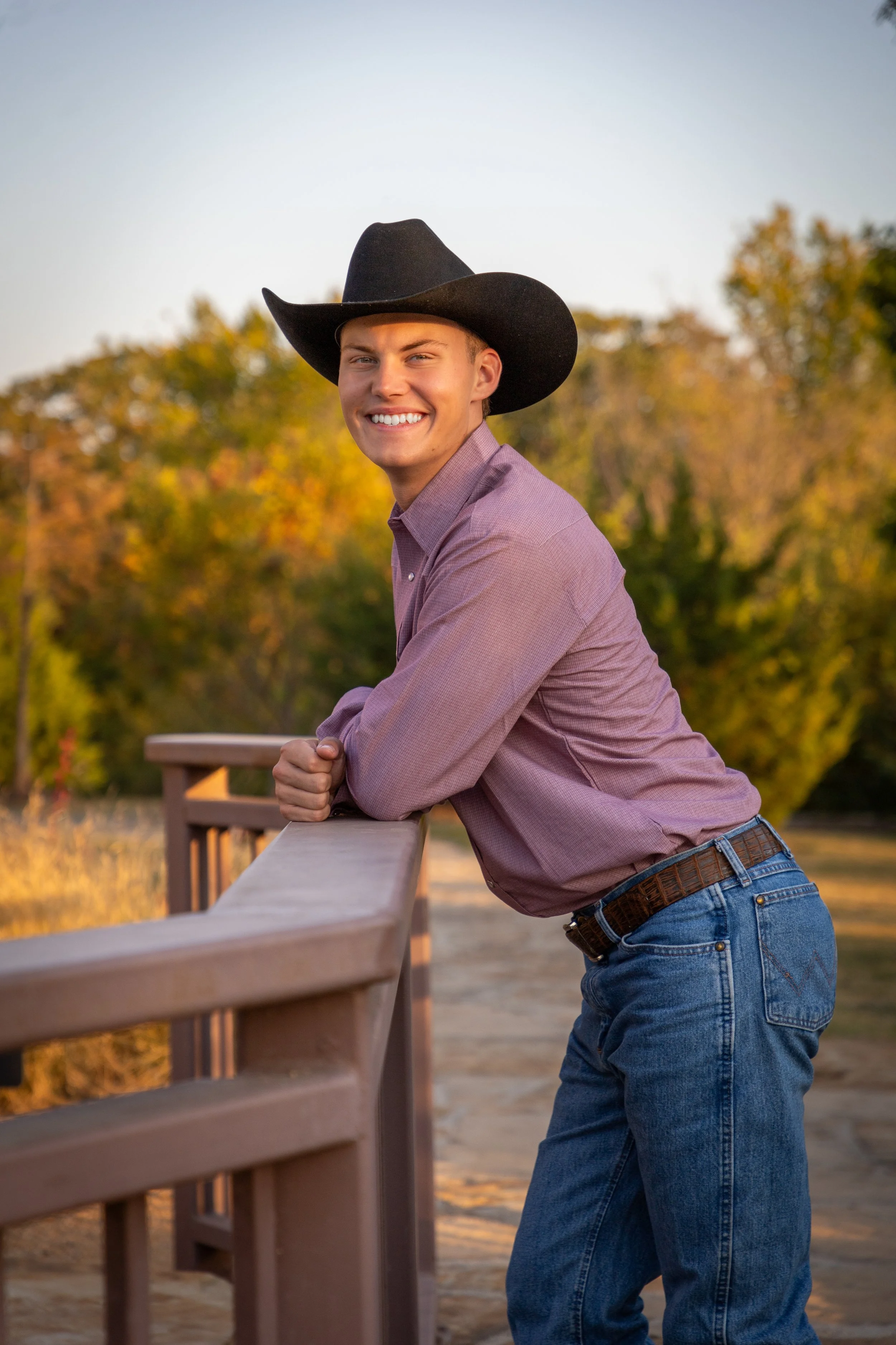 Young man wearing a cowboy hat and purple shirt leaning on a wooden railing outdoors during sunset with trees in the background.