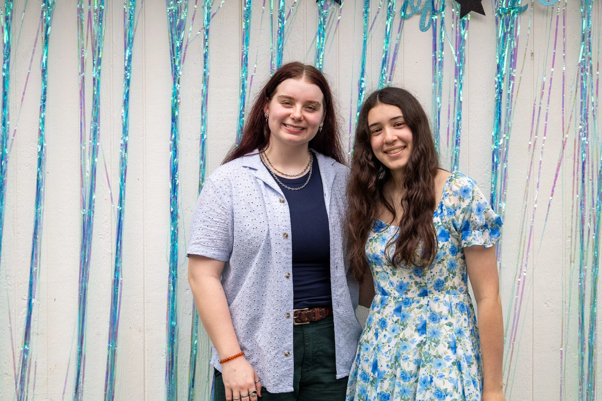 Two young women smiling and standing together in front of a wall decorated with shiny blue and purple streamers.