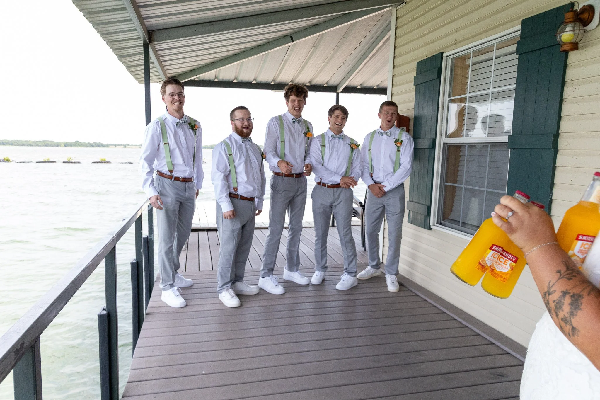 Group of five men dressed in white shirts, gray pants, and suspenders, standing on a dock by the water, smiling and posing for photos during a wedding or celebration.