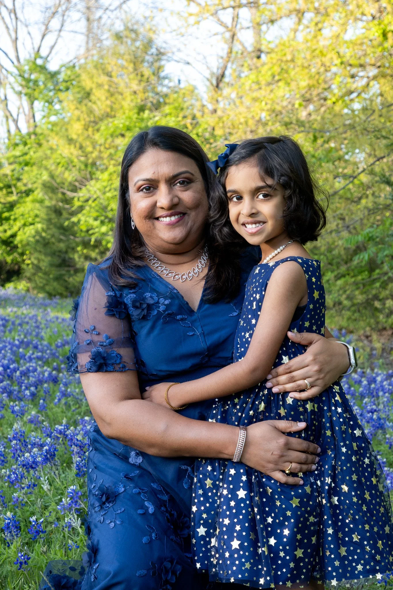 A woman and young girl dressed in matching blue dresses with star patterns, standing outdoors in a field of purple flowers and trees with green leaves.