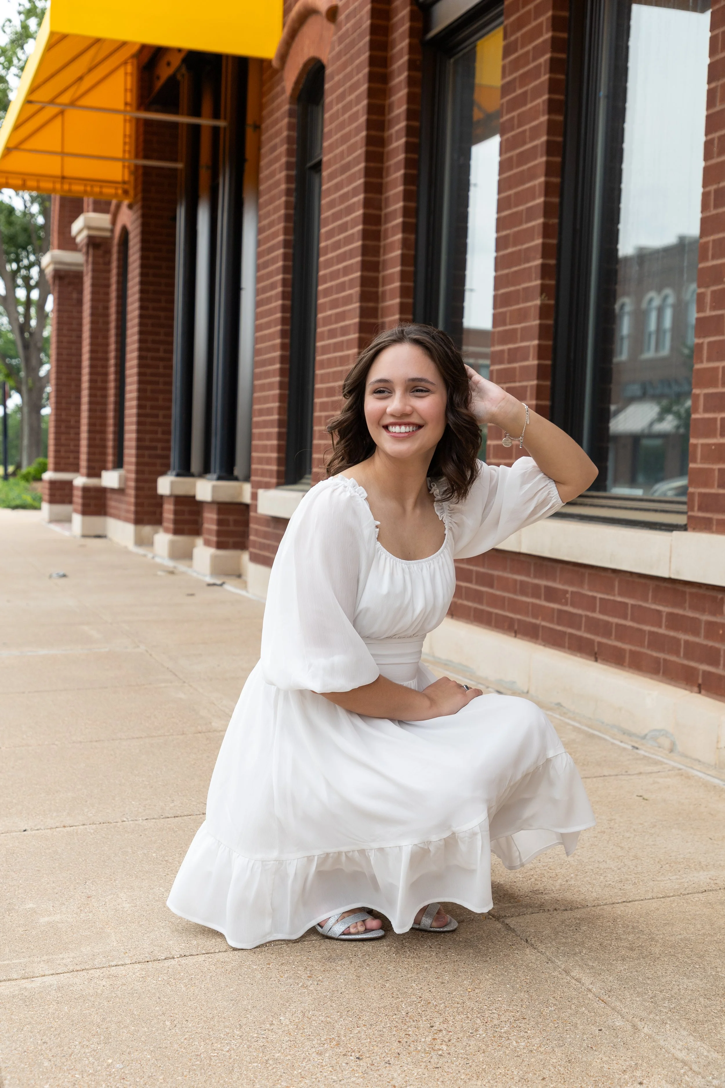 Smiling woman in a white dress crouching on sidewalk in front of brick building with large windows and yellow awning.