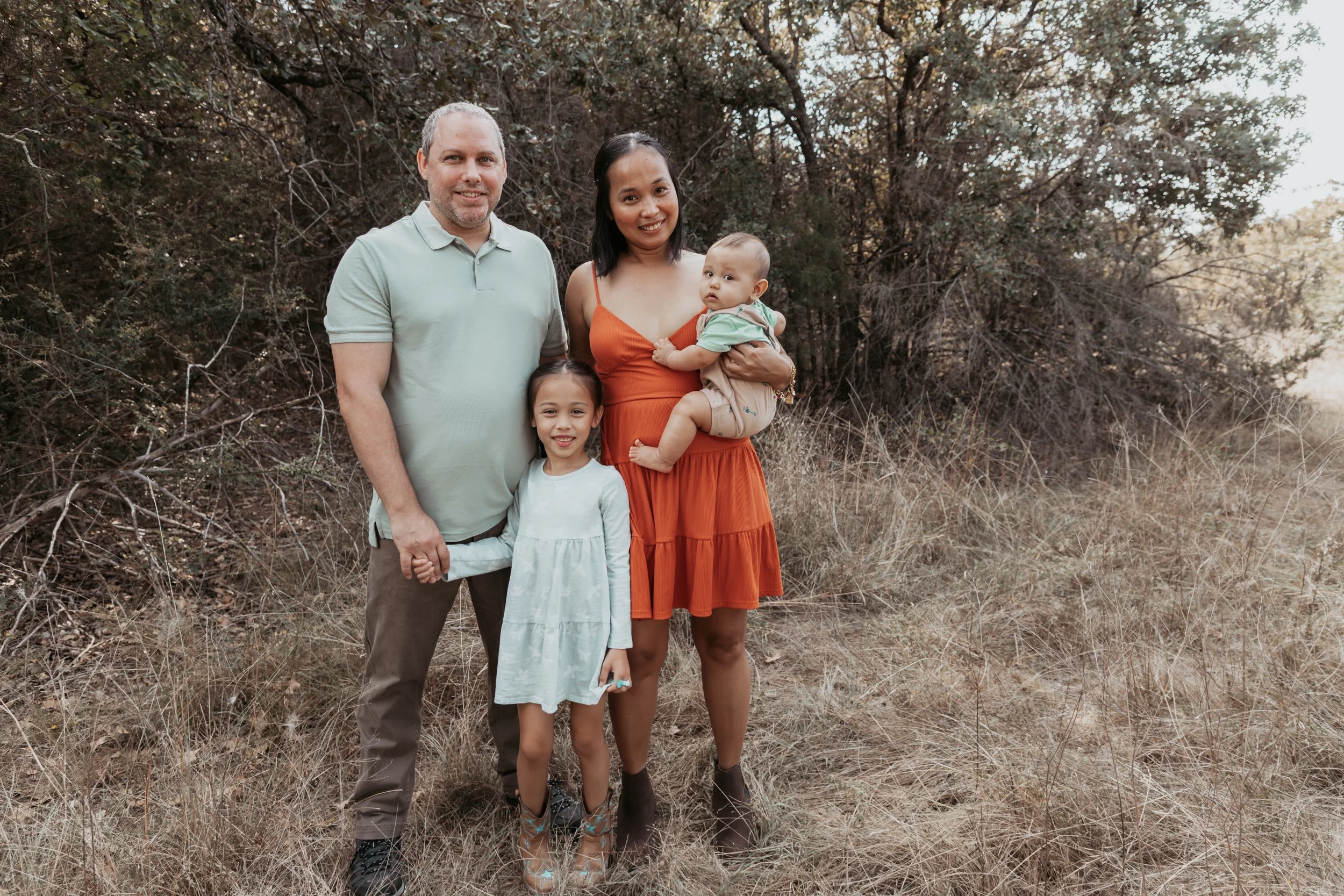 A family of four standing outdoors in a field with trees in the background, holding hands and smiling.