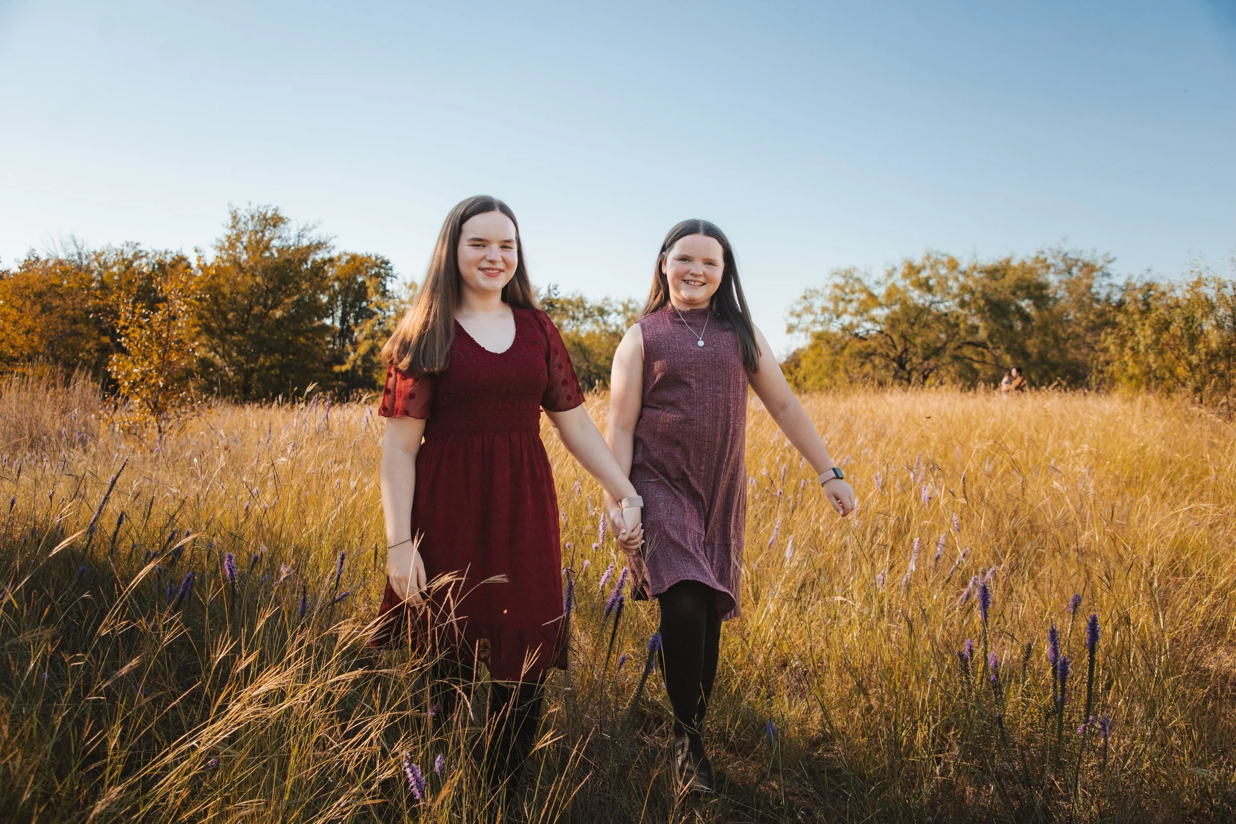 Two young girls holding hands and walking through a grassy field with trees and a blue sky in the background.