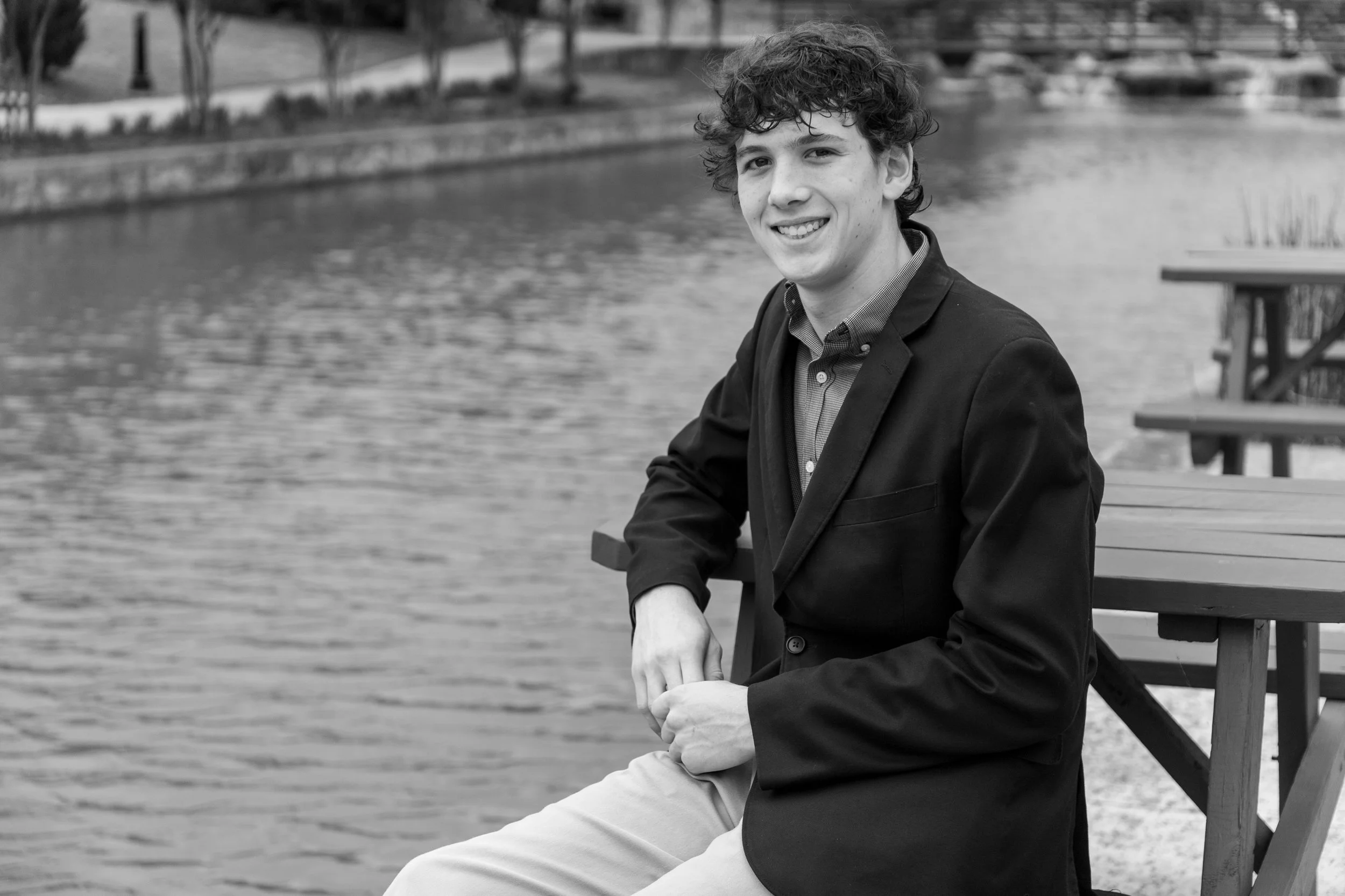 A young man with curly hair smiling and sitting outdoors by a body of water, wearing a dark blazer and light-colored pants.