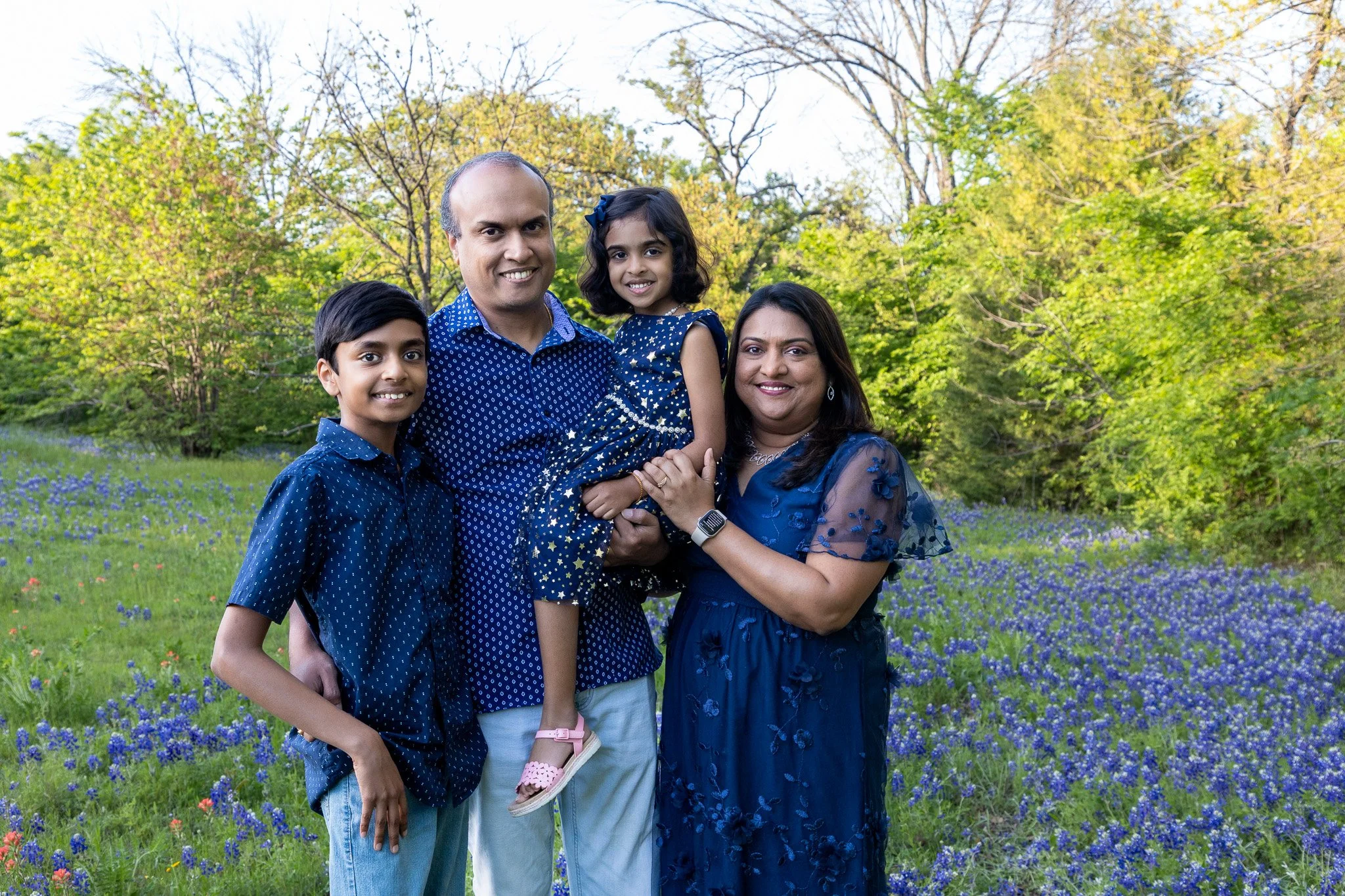 Family of four outdoors in a field of blue flowers with green trees in the background, smiling at the camera.