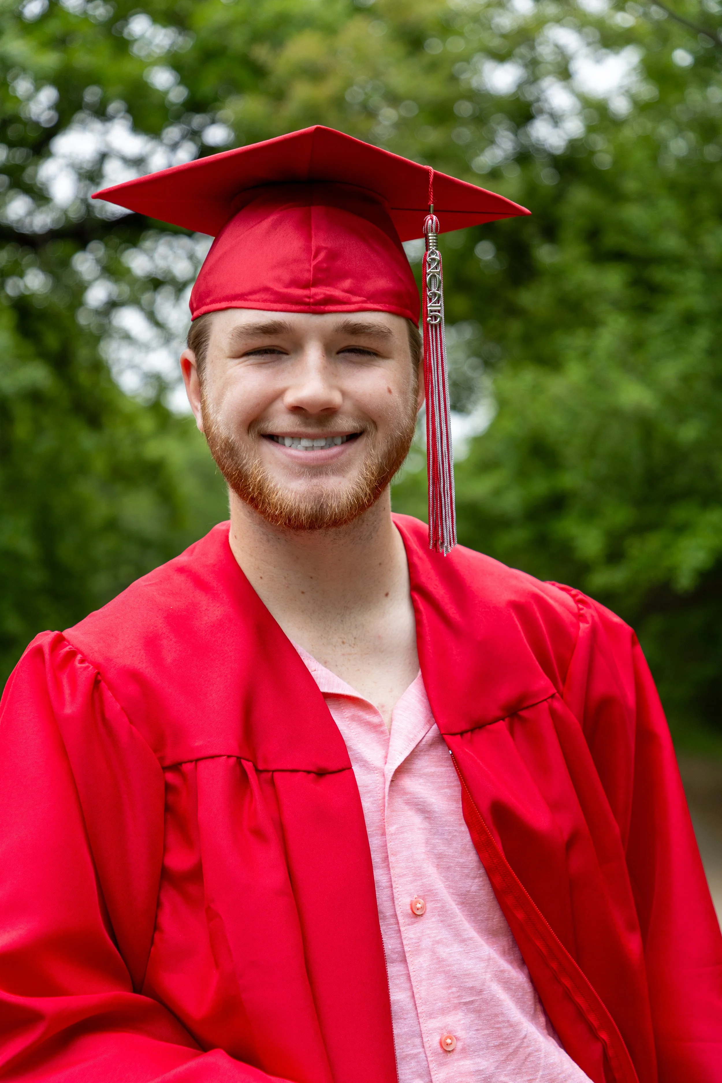 Smiling young man in a red graduation cap and gown outdoors with green trees in the background.