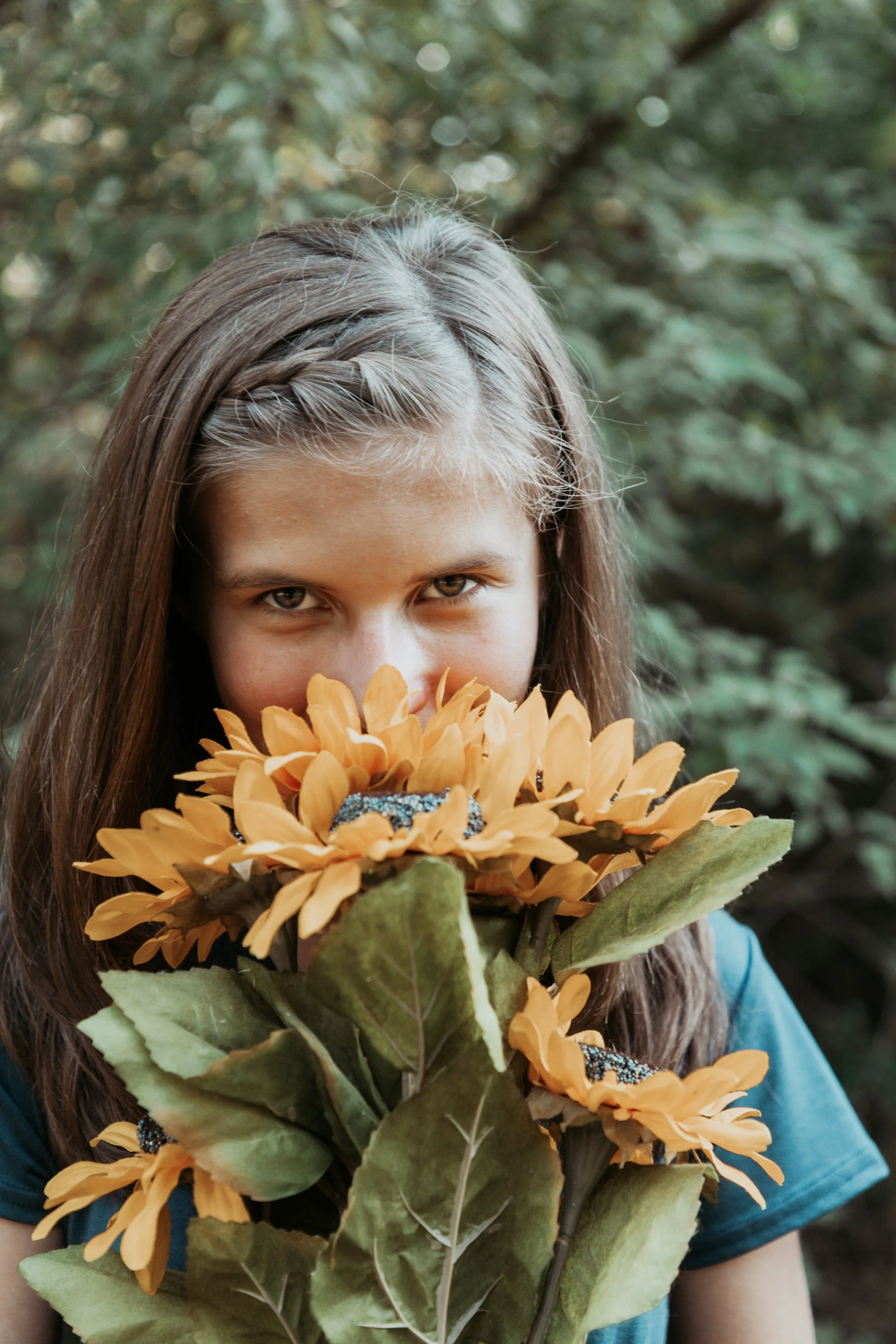 A girl with brown hair peeking over a bunch of yellow flowers outdoors.