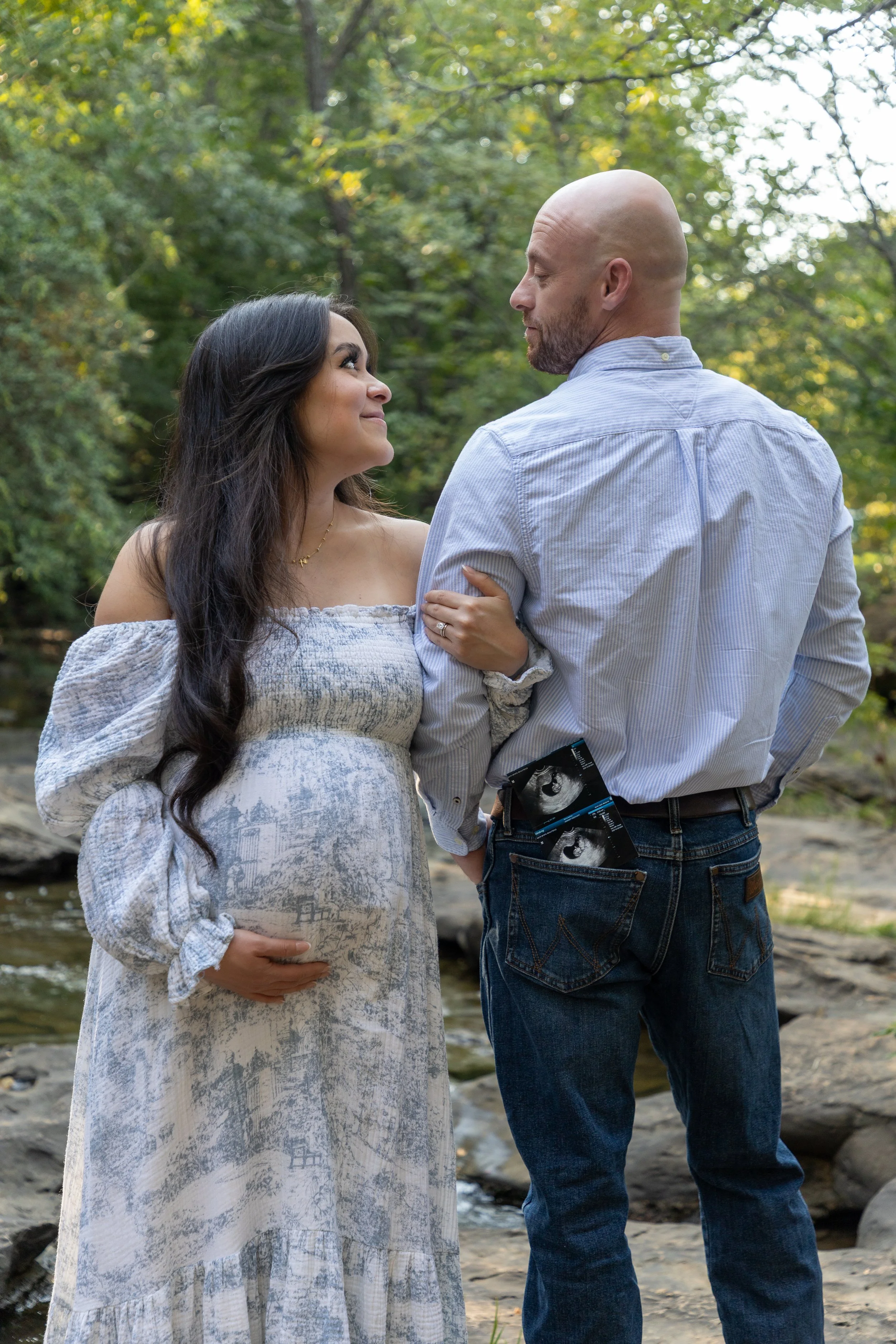 A couple outdoors, the woman is pregnant and holding her belly, while the man has ultrasound pictures tucked into his back pocket. They are standing near a creek with trees in the background, looking at each other lovingly.