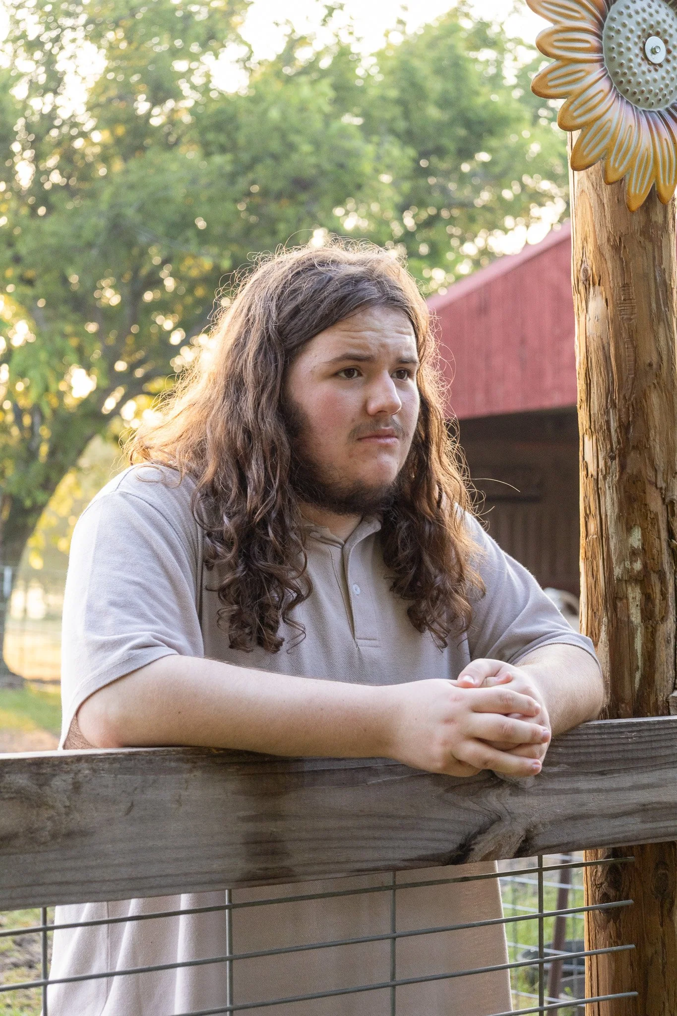 A young man with long, wavy brown hair and a beard, wearing a beige polo shirt, stands outdoors at a wooden fence during the daytime, with trees and a red barn in the background.