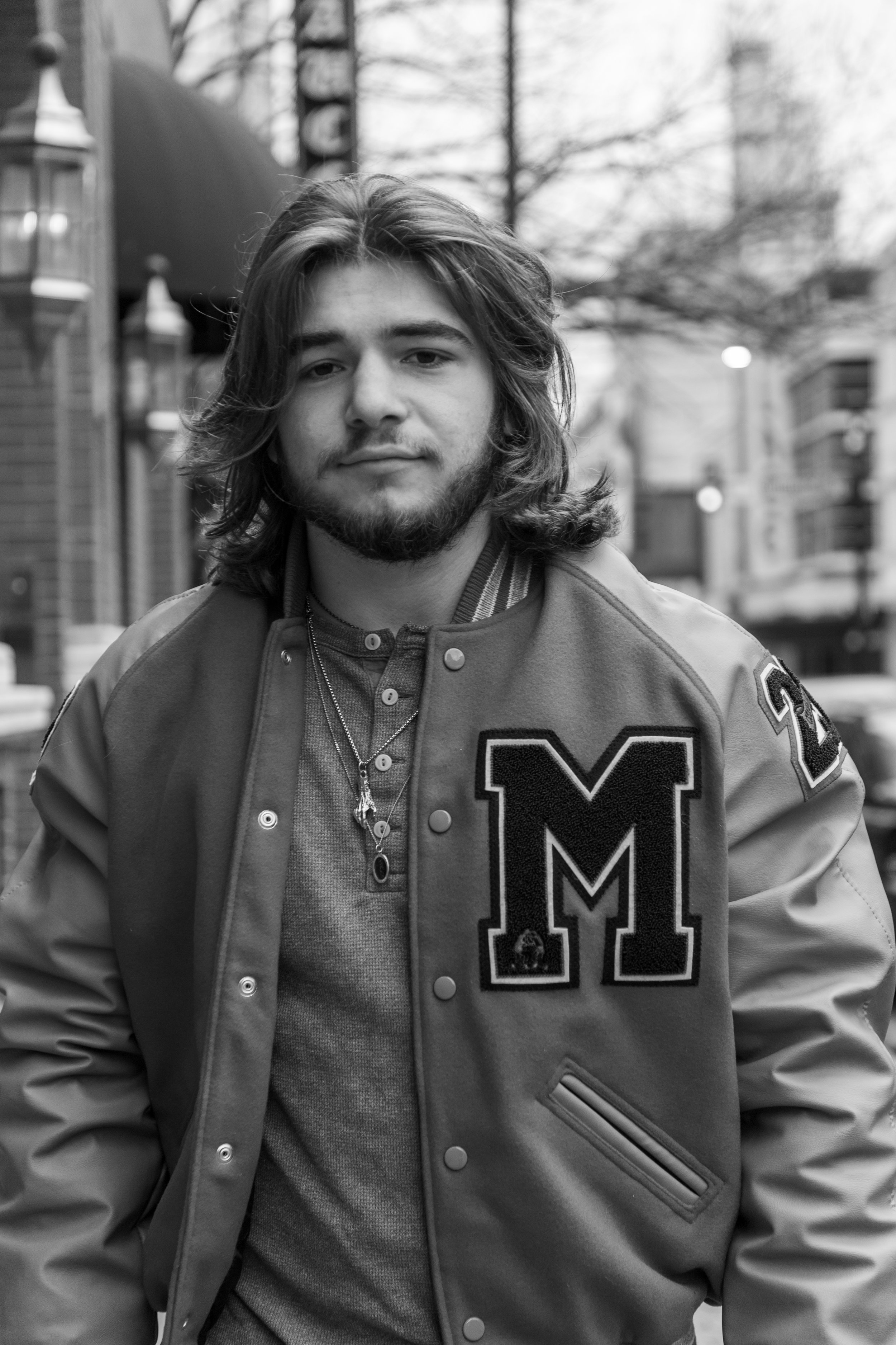 A young man with shoulder-length hair and a beard standing outdoors in an urban environment, wearing a varsity jacket with the letter 'M' on it and layered necklaces.