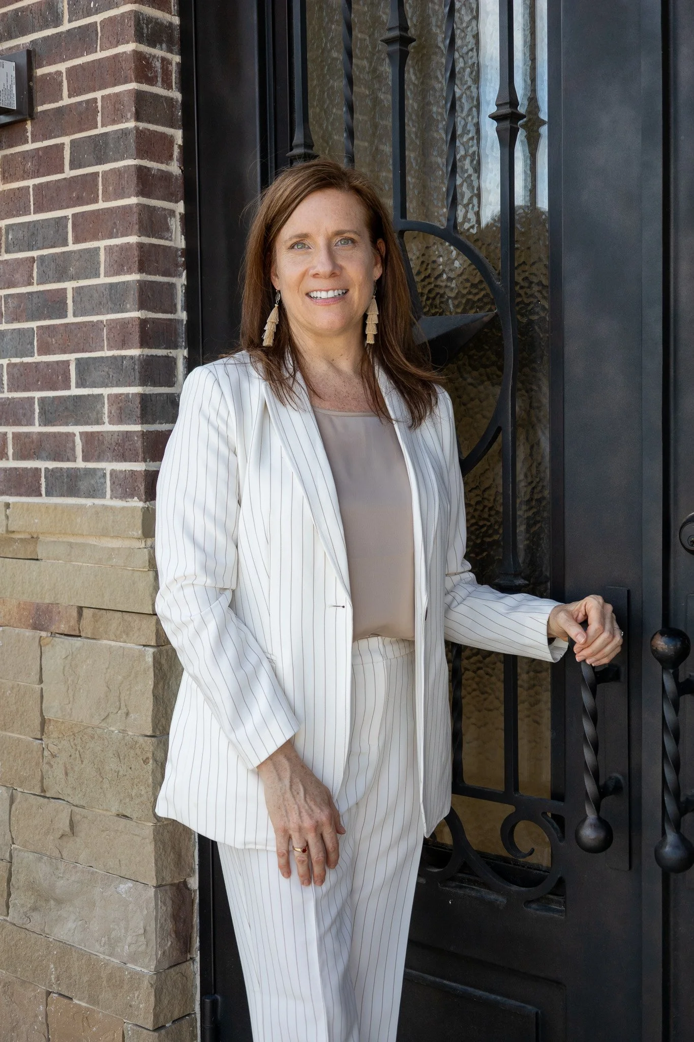 A woman with shoulder-length brown hair wearing a white pinstripe suit standing outside near a black wrought iron gate, holding the gate.