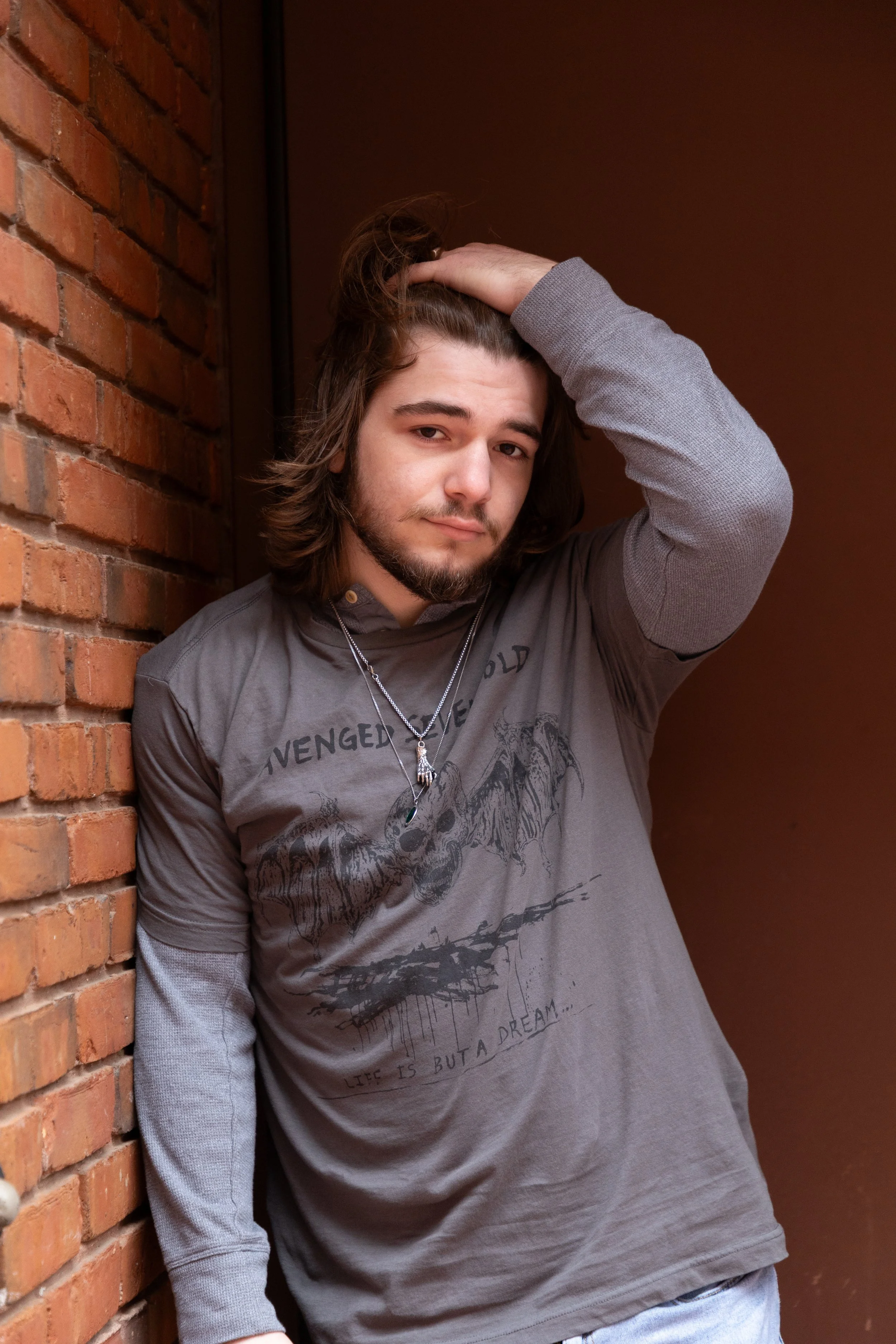 A young man with long hair, a beard, wearing a gray T-shirt with a graphic and text, standing against a brick wall and a painted wall, looking at the camera with a relaxed expression.