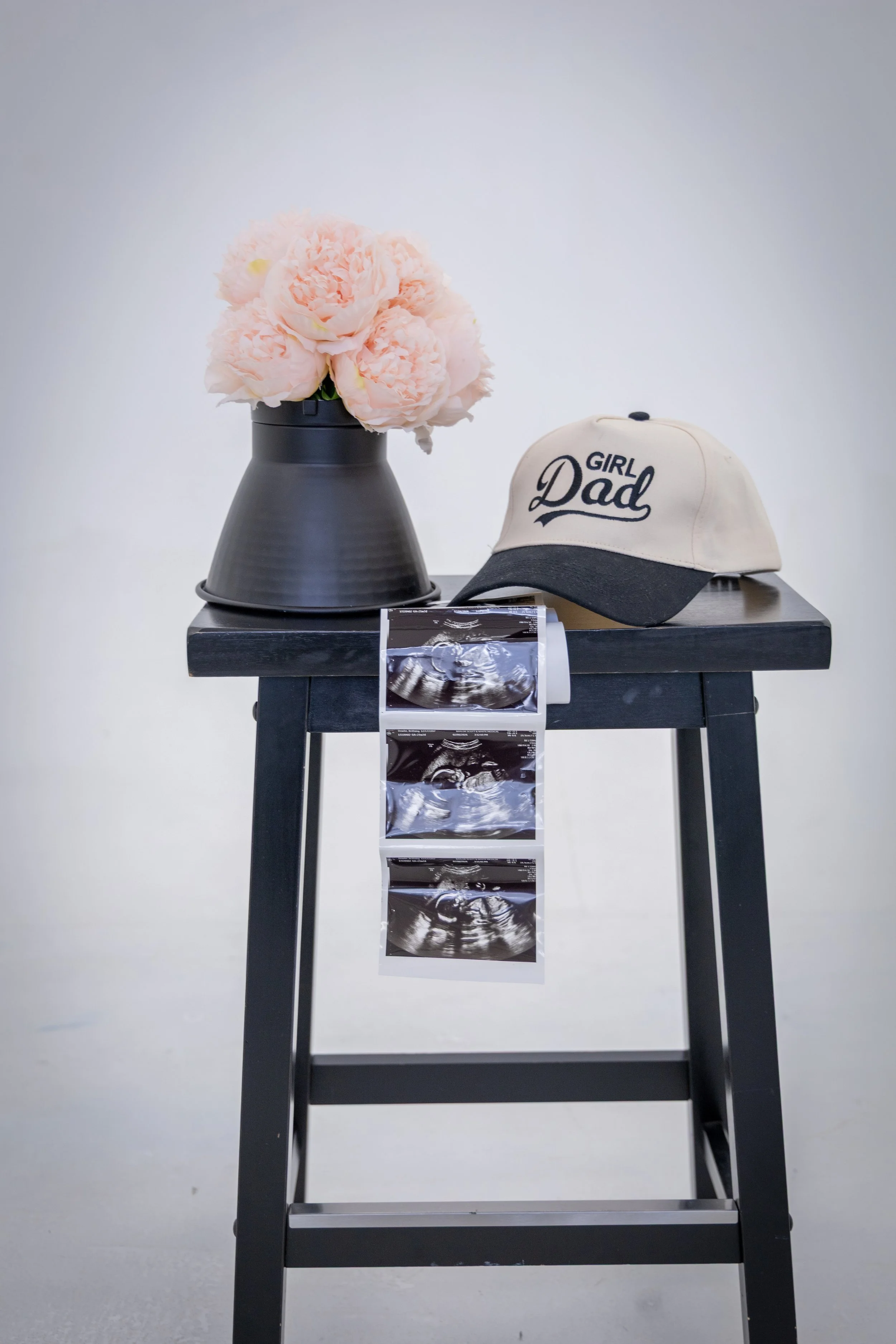 A black table with light pink peony flowers in a dark vase on the left, a beige baseball cap with black text reading 'GIRL Dad' on the right, and four ultrasound photos hanging from the edge of the table. The background is plain white.