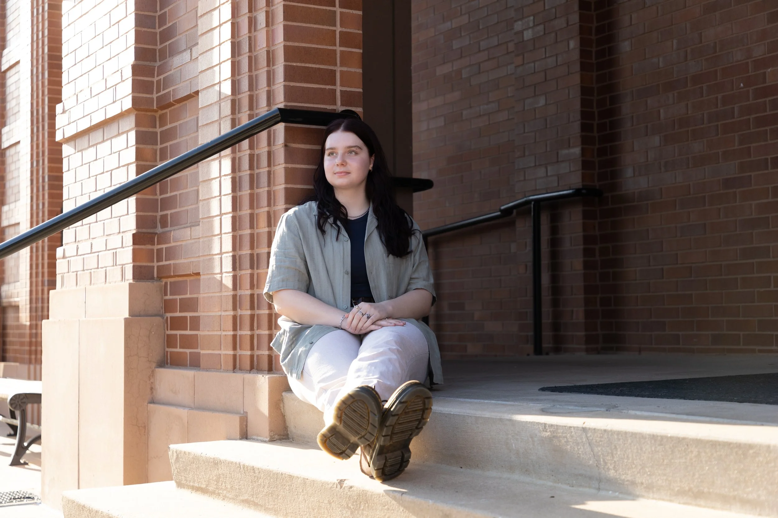 A young woman with black hair sitting on steps outside a brick building, looking to the side.
