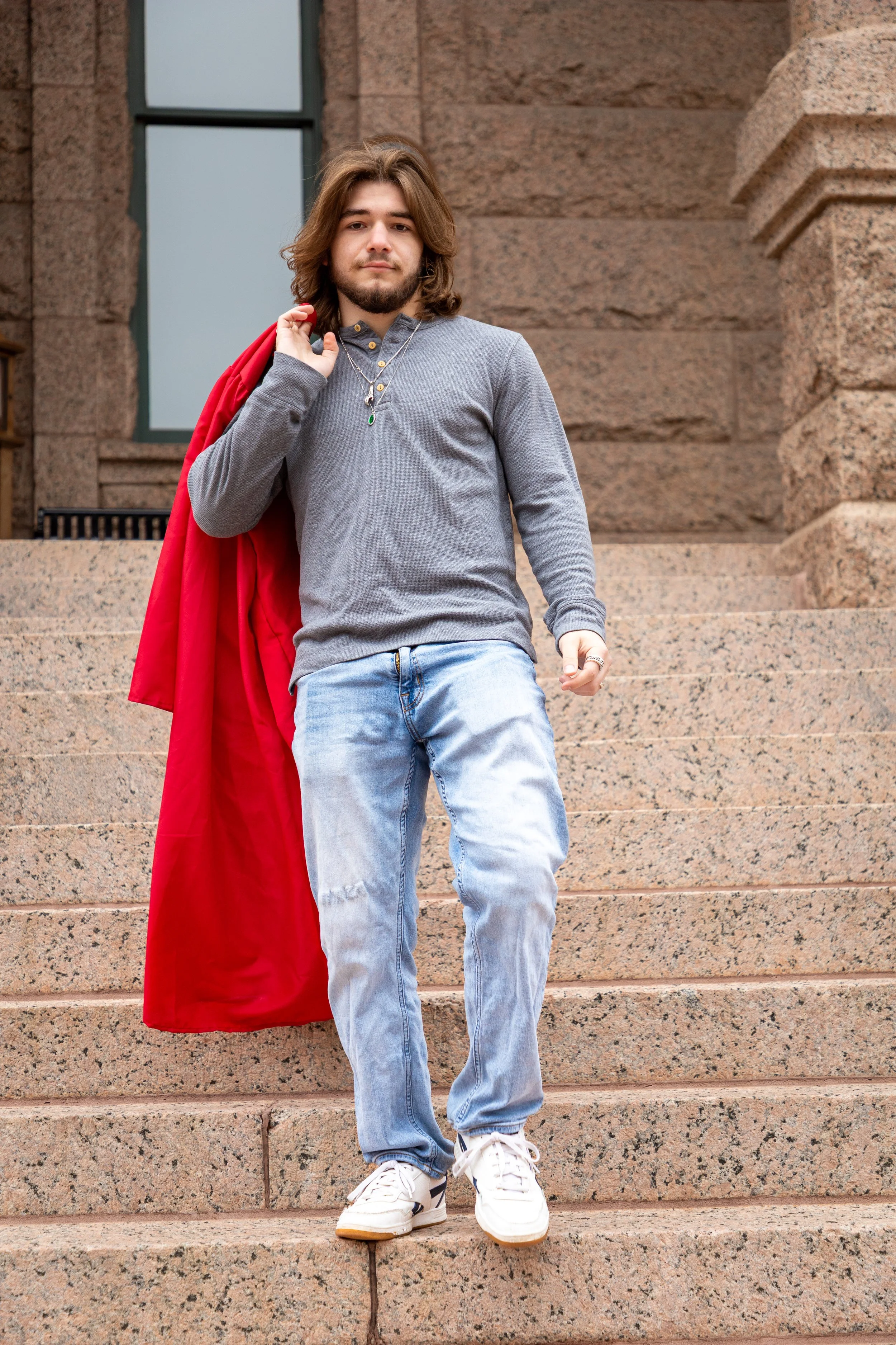 A young man with shoulder-length hair and a beard standing on stone steps outside a building, carrying a red coat over his shoulder, wearing a gray sweater, light blue jeans, and white sneakers.