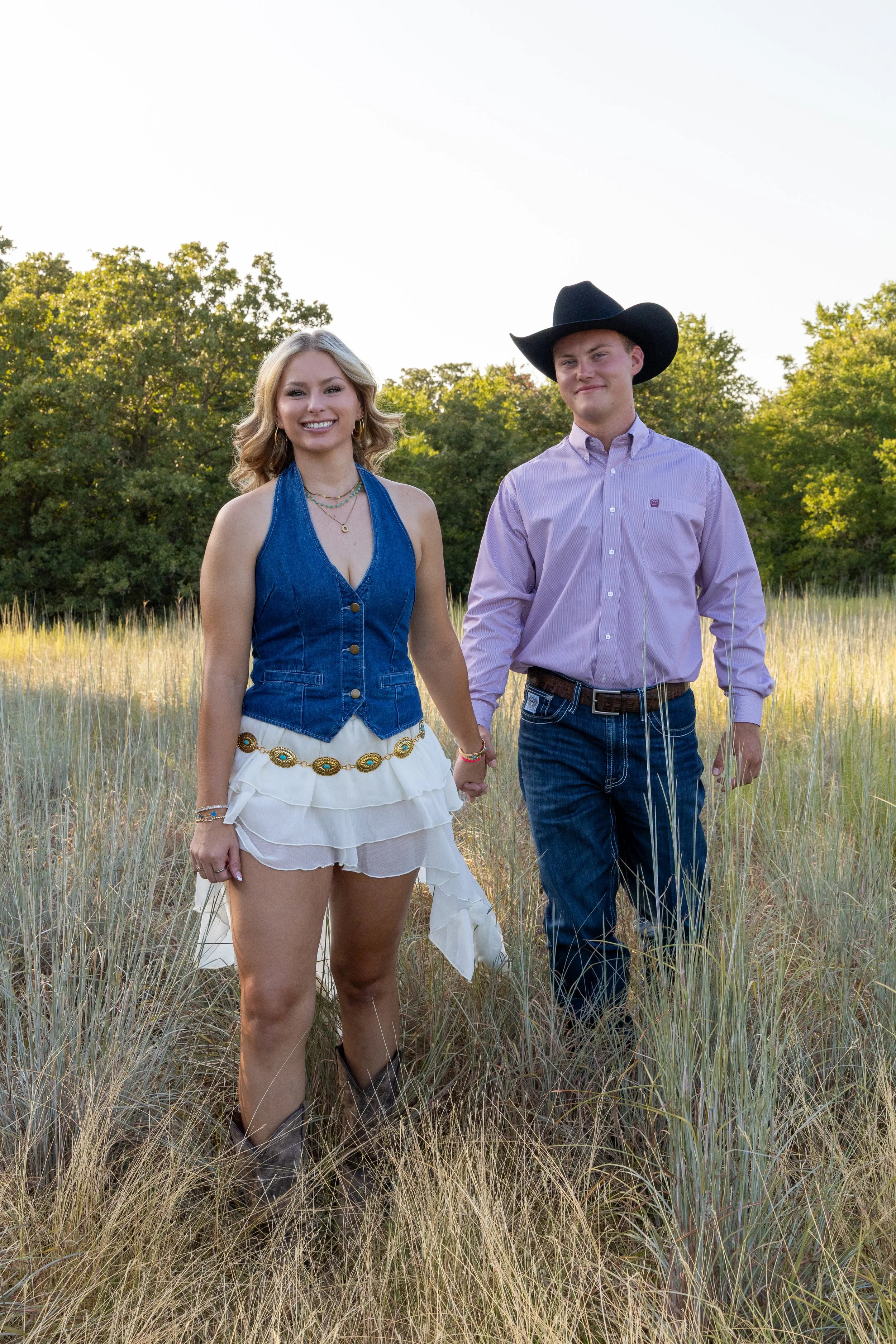 A young woman and a young man holding hands and walking in a grassy field with trees in the background during daytime. The woman is smiling, wearing a blue denim vest over a white dress, and boots. The man is wearing a cowboy hat and a light purple b