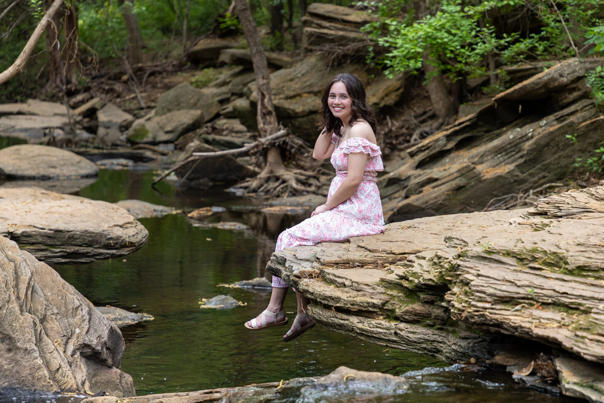 A smiling young woman in a pink floral dress sits on a large flat rock by a small creek in a wooded area, with her feet resting just above the water.