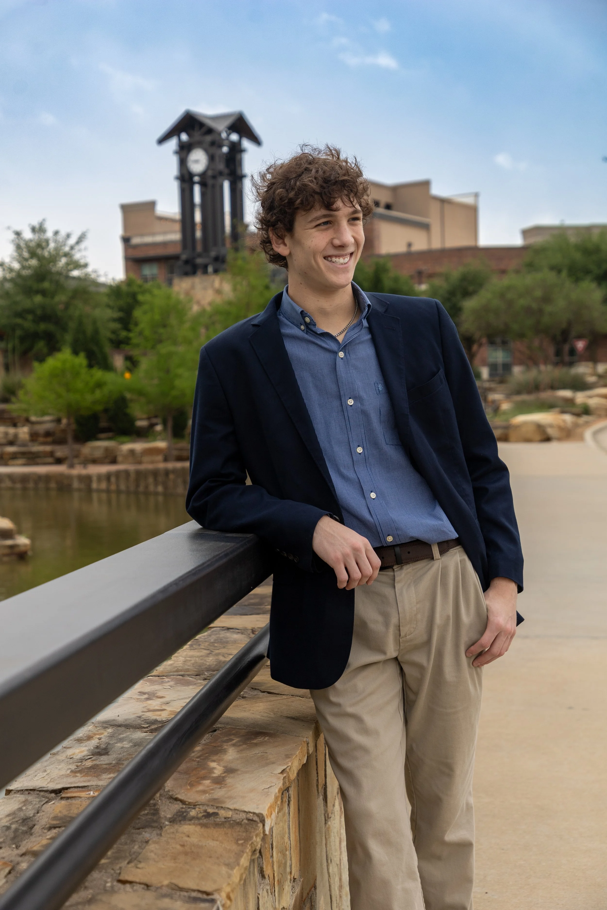 A young man with curly brown hair, wearing a navy blazer, blue dress shirt, and khaki pants, leaning against a stone railing outdoors near a lake with trees and a building with a clock tower in the background.