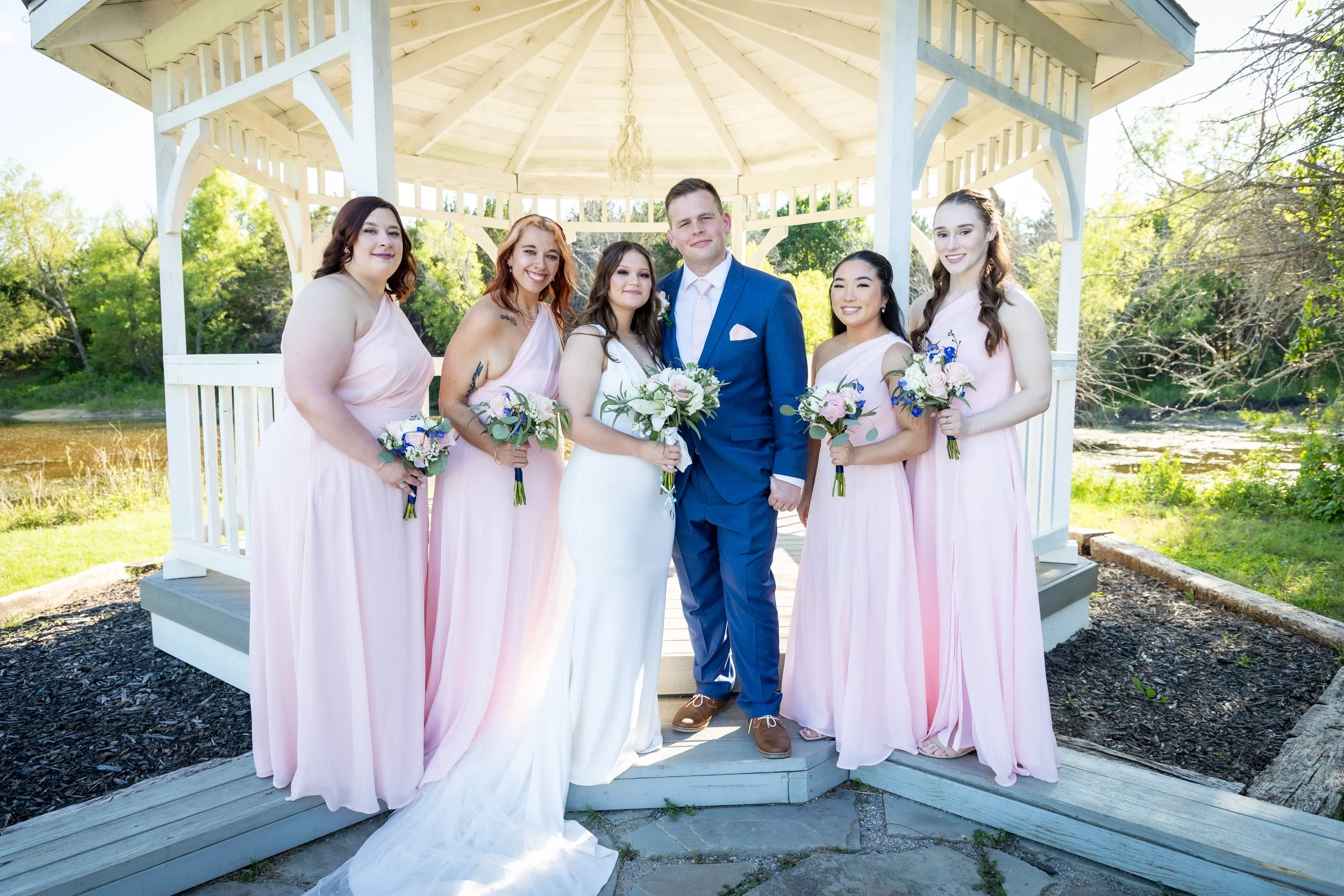 A wedding party of six people standing under a white gazebo outdoors, with the bride in a white gown holding a bouquet, the groom in a blue suit, and four bridesmaids in pink dresses holding bouquets.