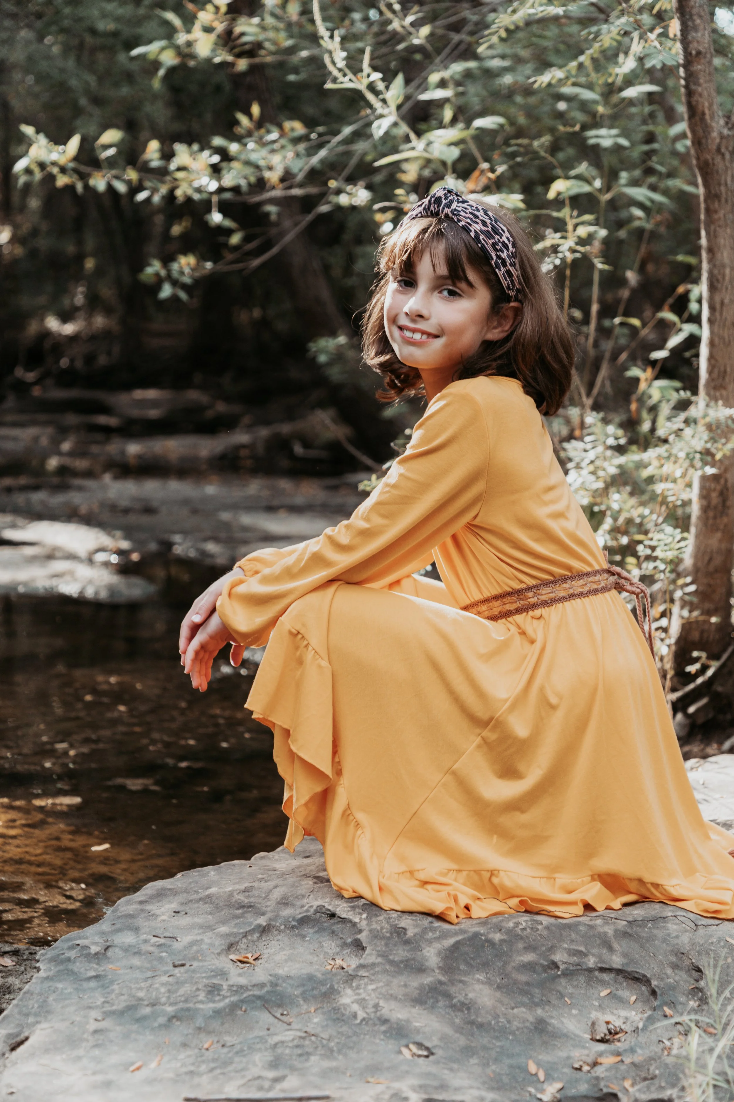 A young girl sitting on a rock near a creek in a wooded area, smiling at the camera. She is wearing a yellow dress and a patterned headband.