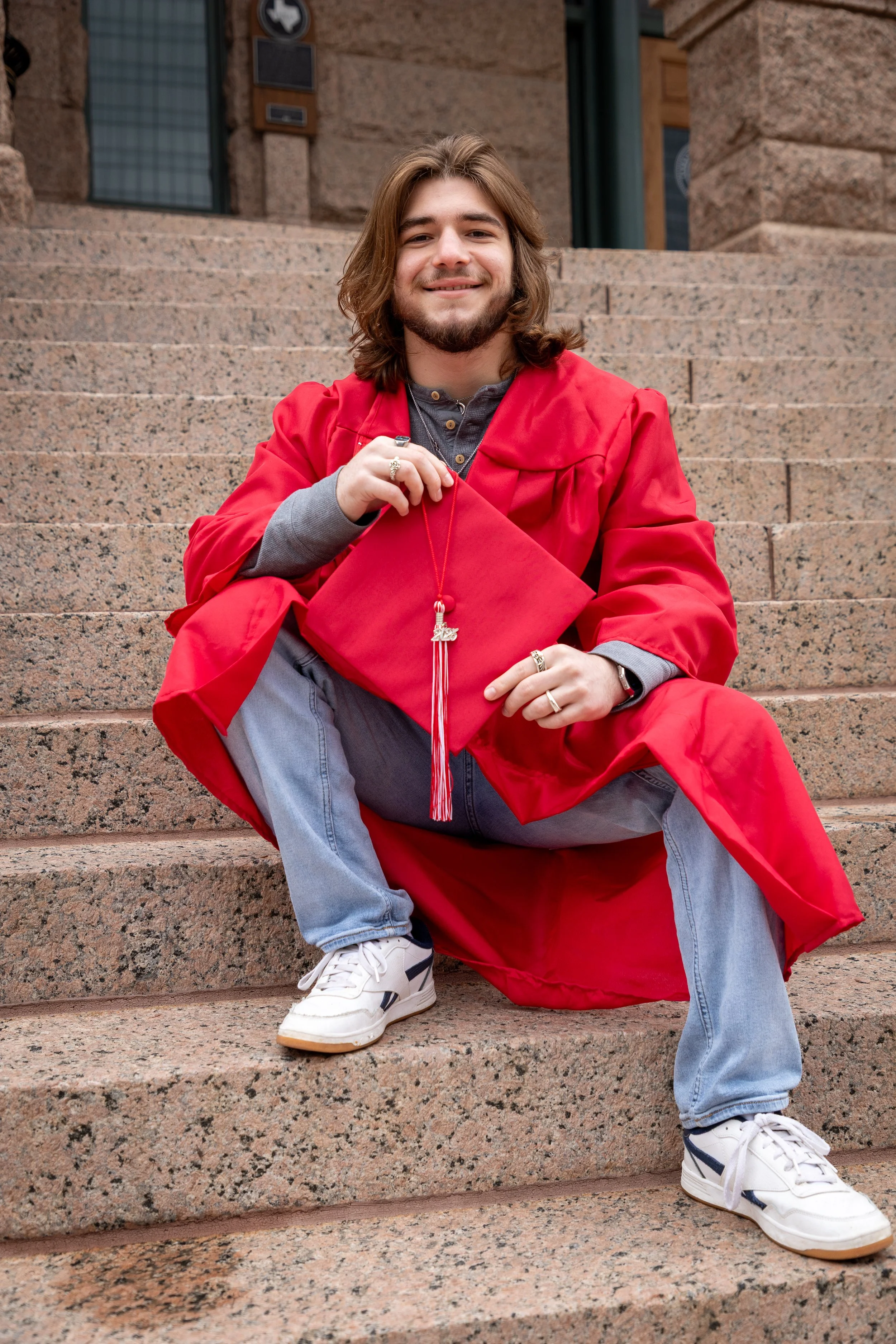 A young man in a red graduation gown and cap, sitting on stone steps outside a building, holding his cap in front of him, smiling.