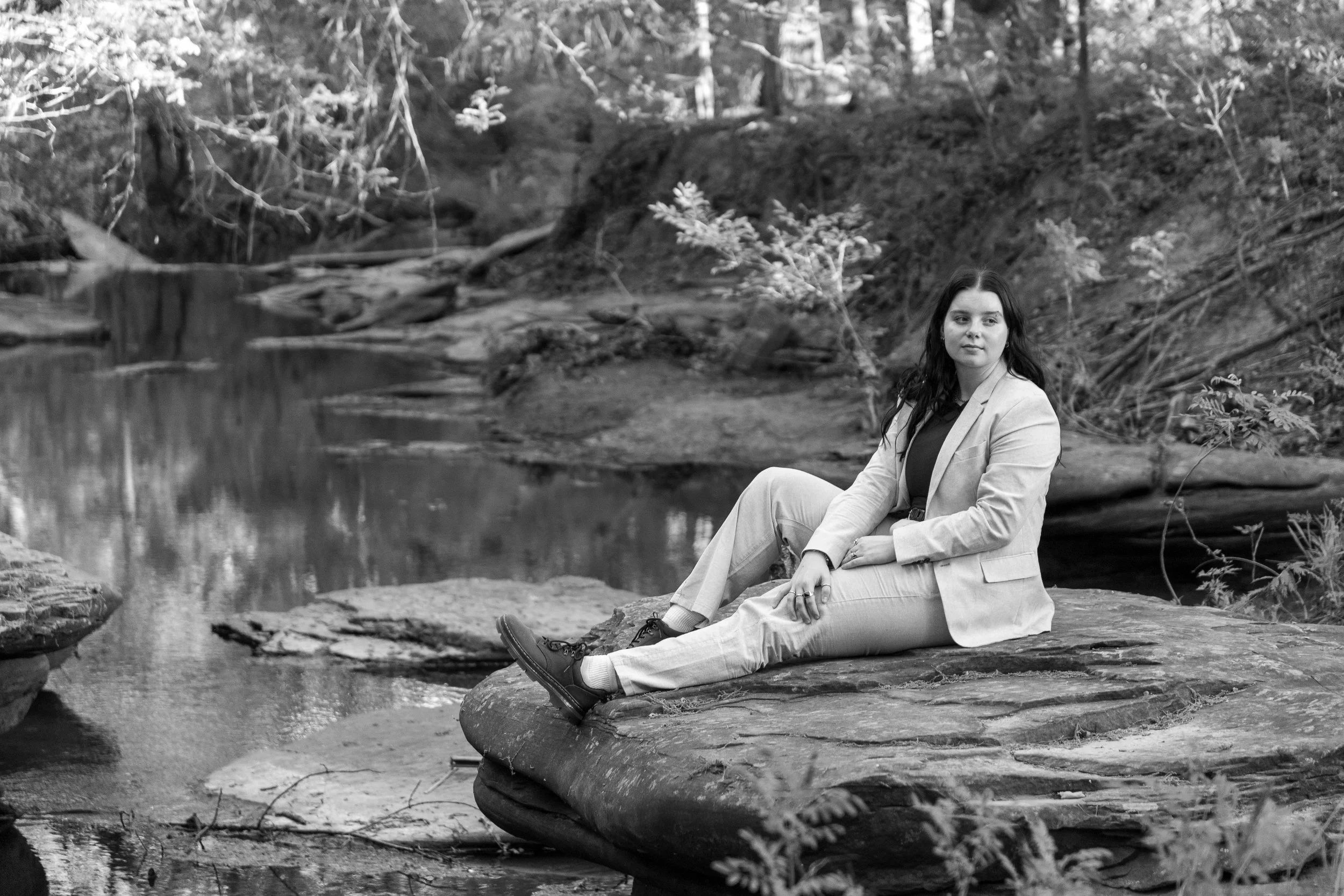 A woman dressed in a suit sitting on a large rock by a stream in a forested area