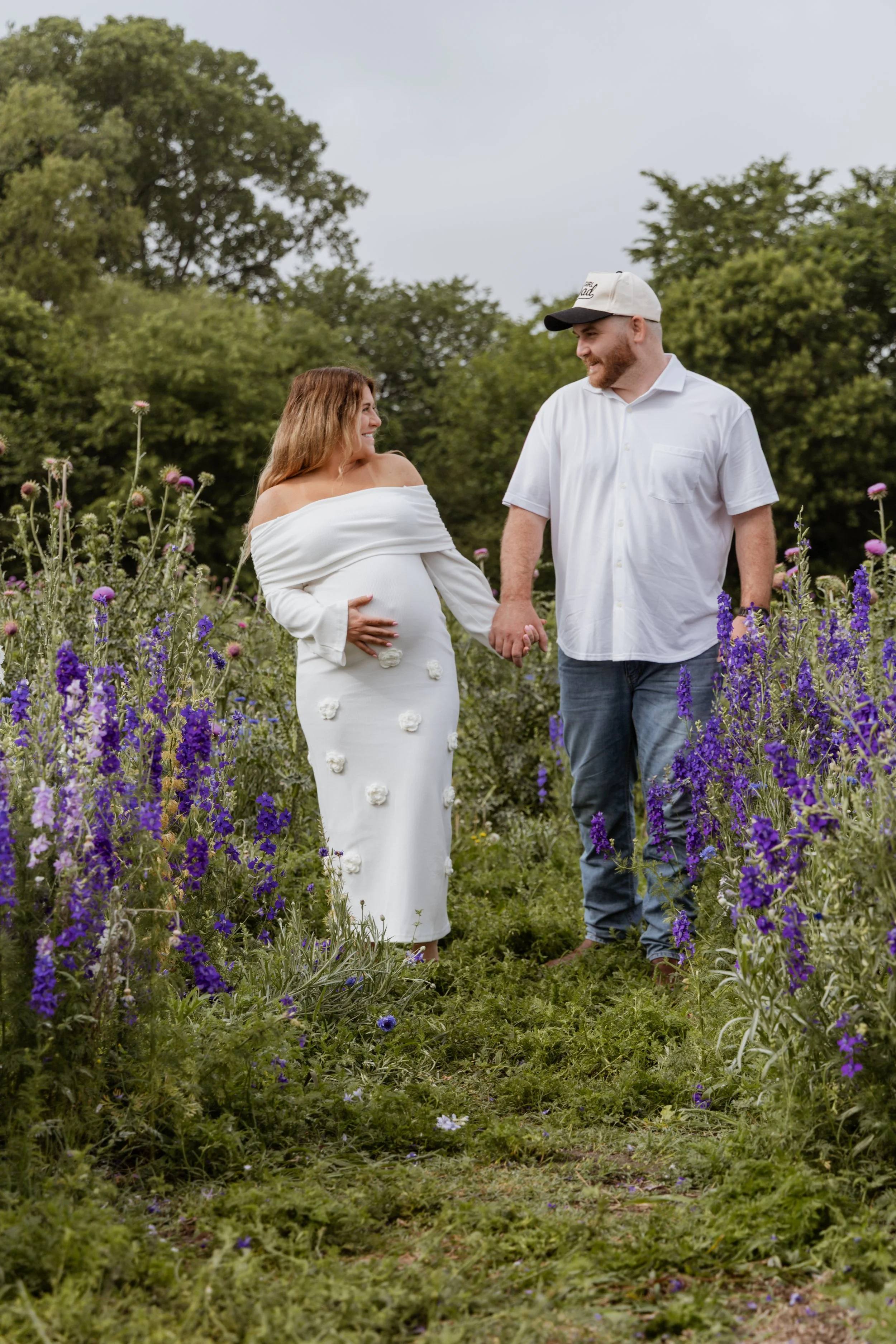 A pregnant woman in a white off-shoulder dress with floral embellishments holds hands with a man in a white shirt and jeans, walking through a field of purple and pink flowers with green trees in the background.