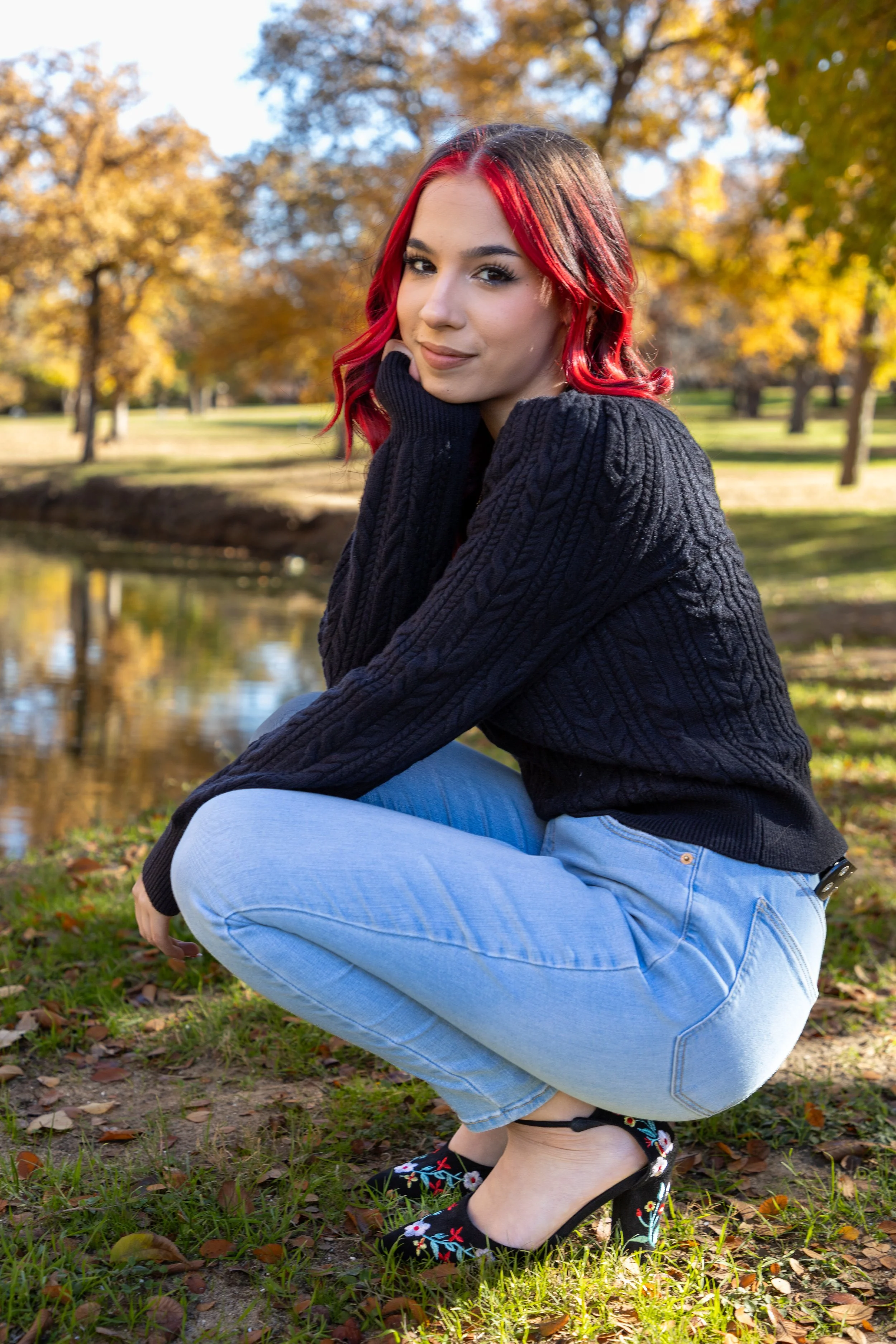 A young woman with bright red hair, wearing a black cable-knit sweater, light blue jeans, and black embroidered high heels, squatting by a pond in a park with autumn trees in the background.