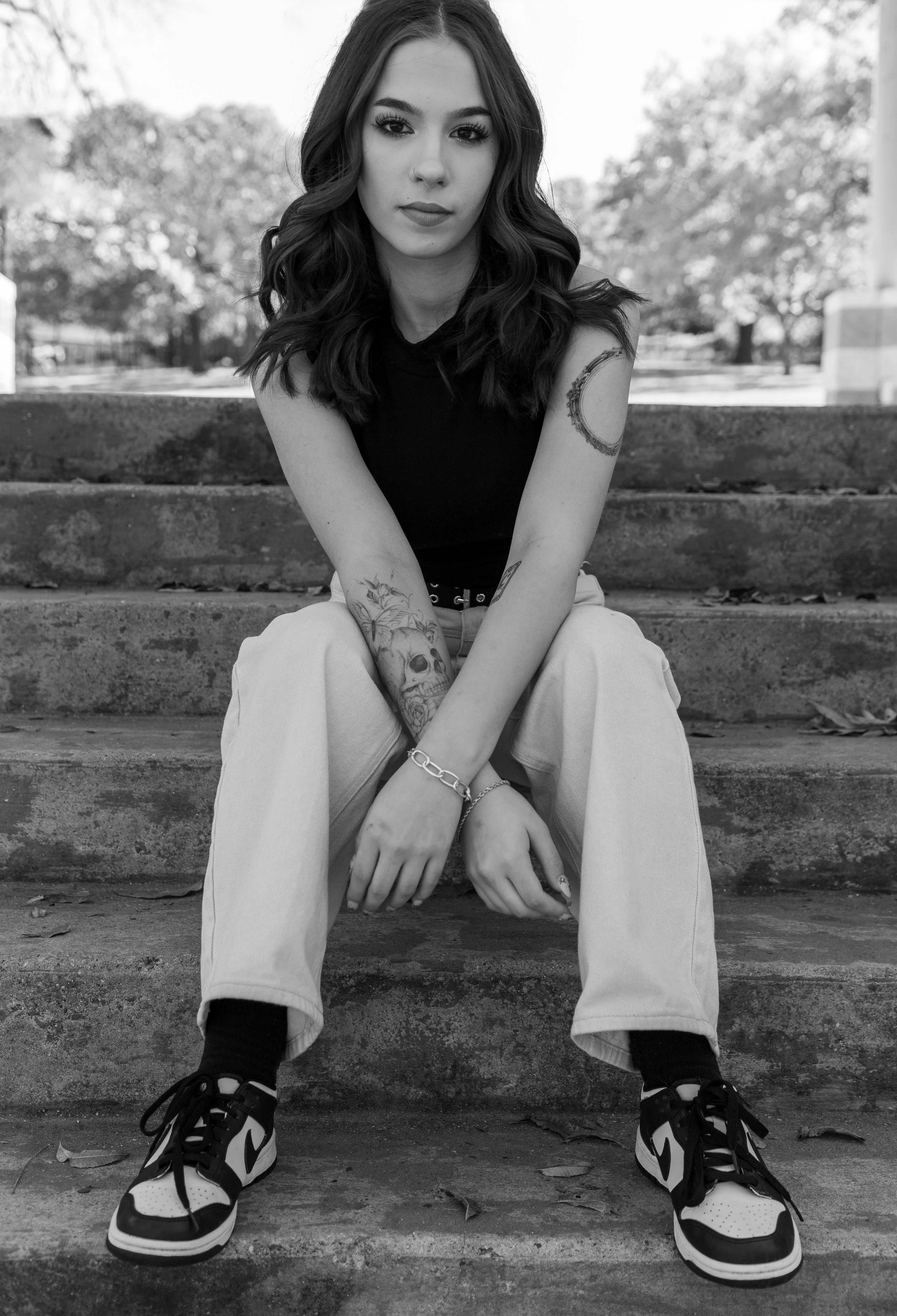 A young woman with long wavy hair sitting on outdoor stone steps, wearing a black sleeveless top, light-colored pants, sneakers, and jewelry. She has visible tattoos on her arms and a nose piercing, with a calm expression and trees in the background.