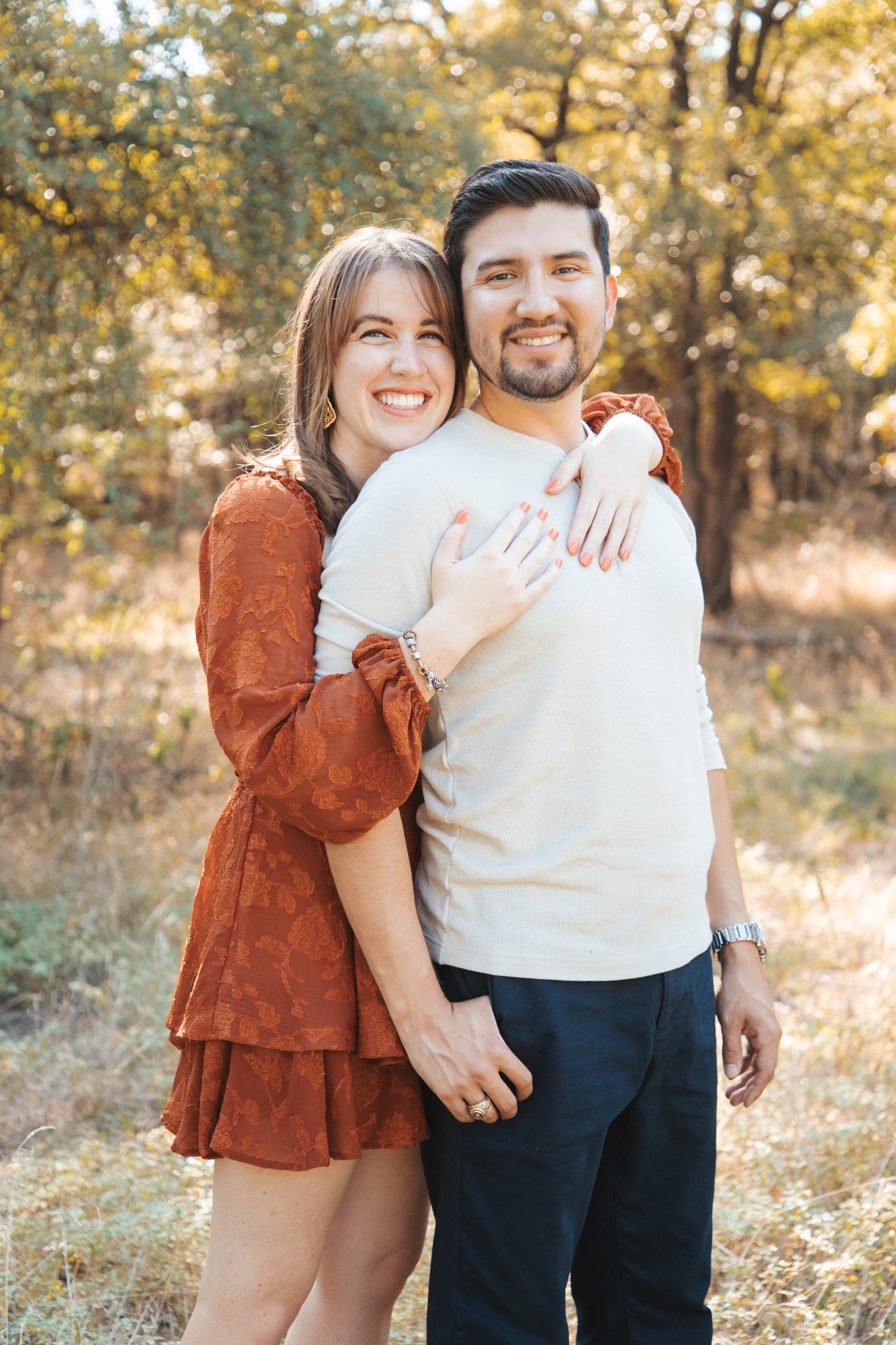 A happy couple stands outdoors on a sunny fall day, with trees and golden foliage in the background. The woman has shoulder-length brown hair and is wearing a rust-colored dress with a floral pattern, while the man has short dark hair, a beard, and i