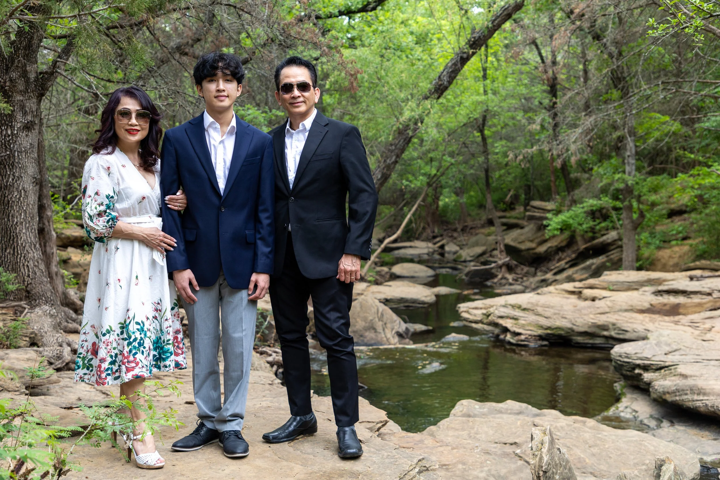 Family of three standing on rocks beside a creek in a wooded area, dressed in semi-formal attire, with the woman in a white dress with floral patterns, the young man in a blue blazer and gray pants, and the man in a black suit and sunglasses.