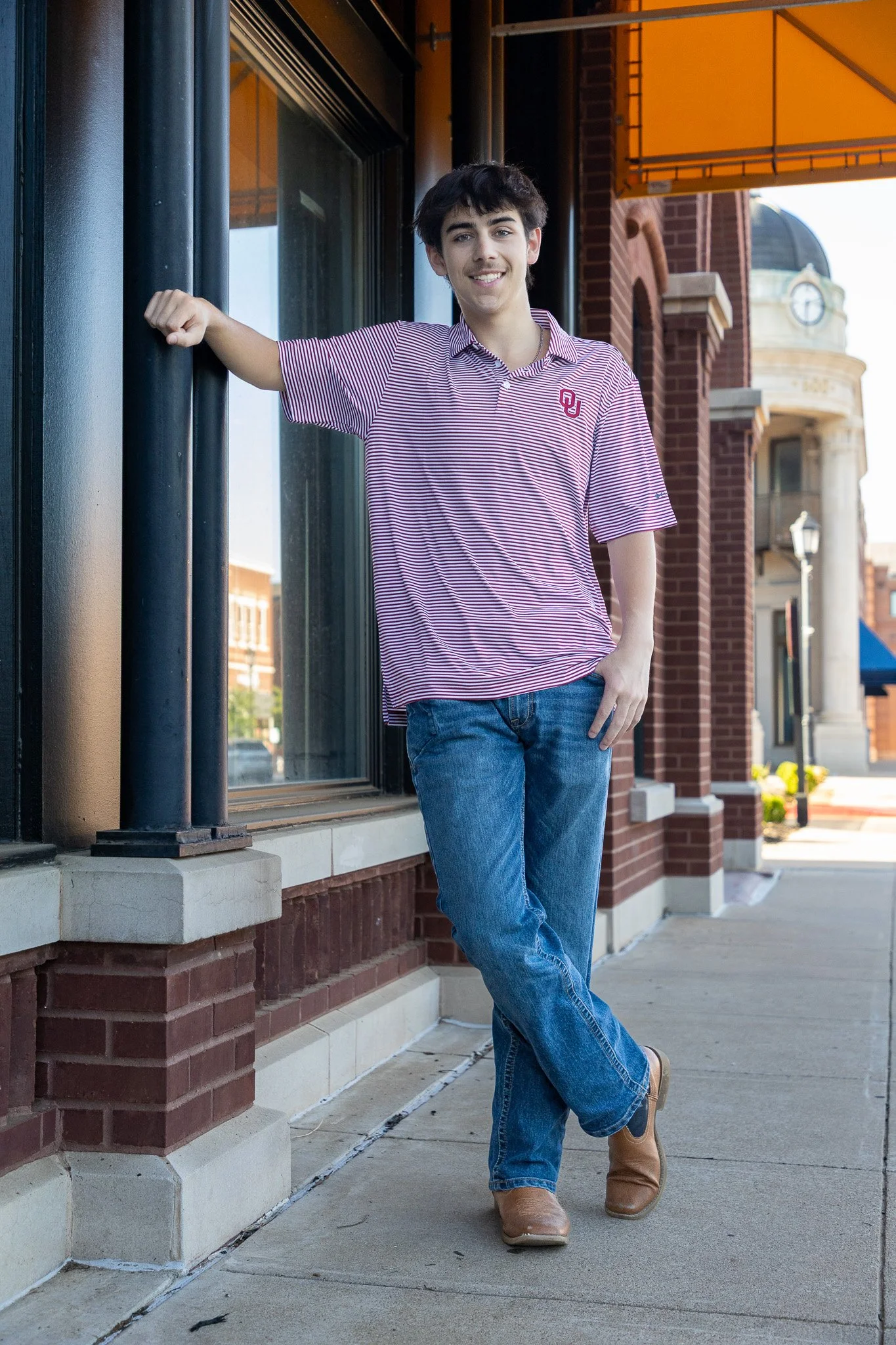 A young man with dark hair, wearing a striped polo shirt, jeans, and brown boots, standing outside on a sidewalk, leaning against a building with a glass window and brick facade, smiling at the camera.