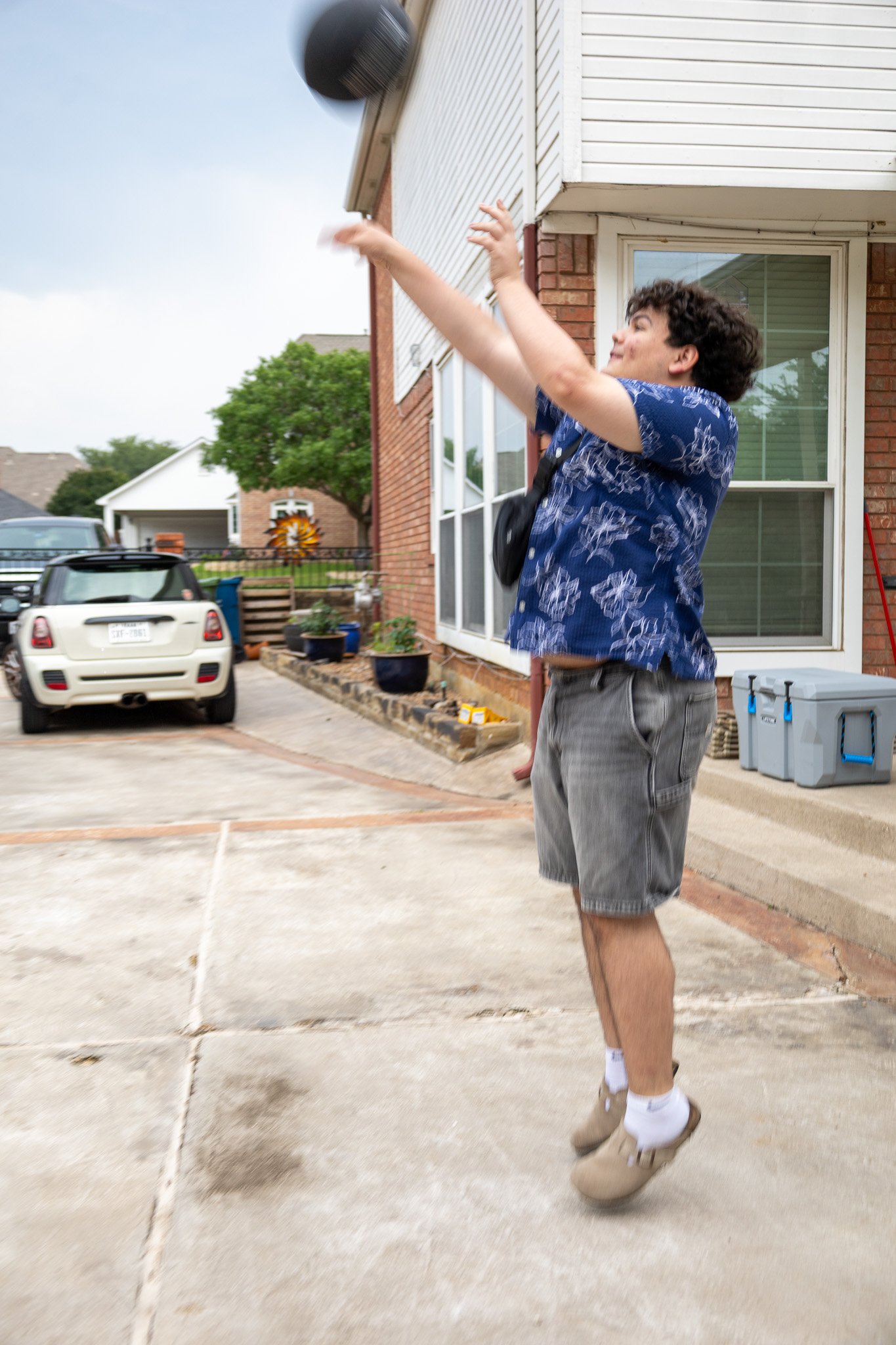 A young man with curly hair, wearing a blue floral shirt, gray shorts, and tan shoes, is playing volleyball outdoors on a concrete driveway beside a house. He is jumping to hit a black volleyball with his right hand.
